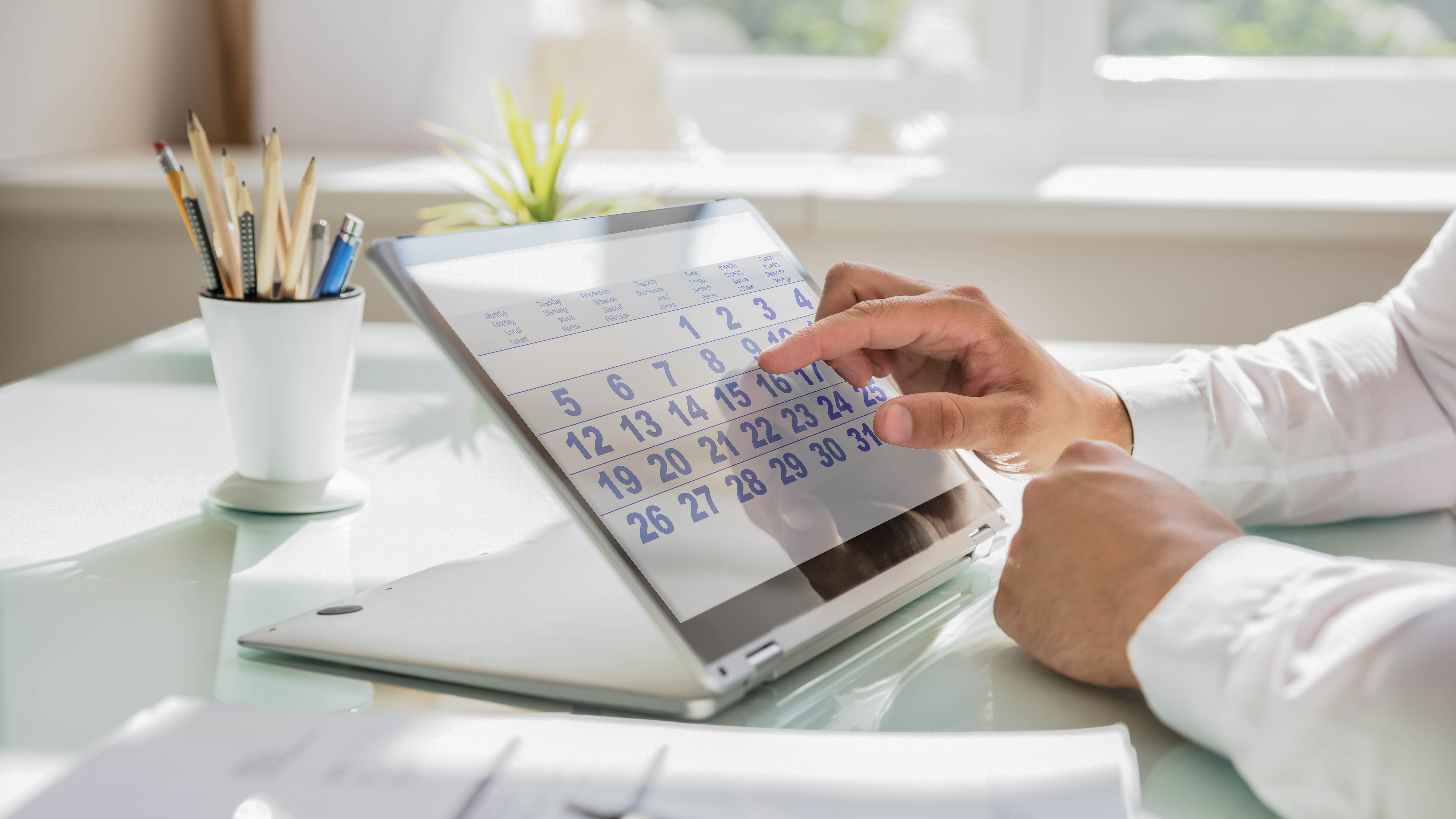 Eine Hand tippt auf einen digitalen Kalender auf einem Tablet, daneben ein Topf mit Stiften.