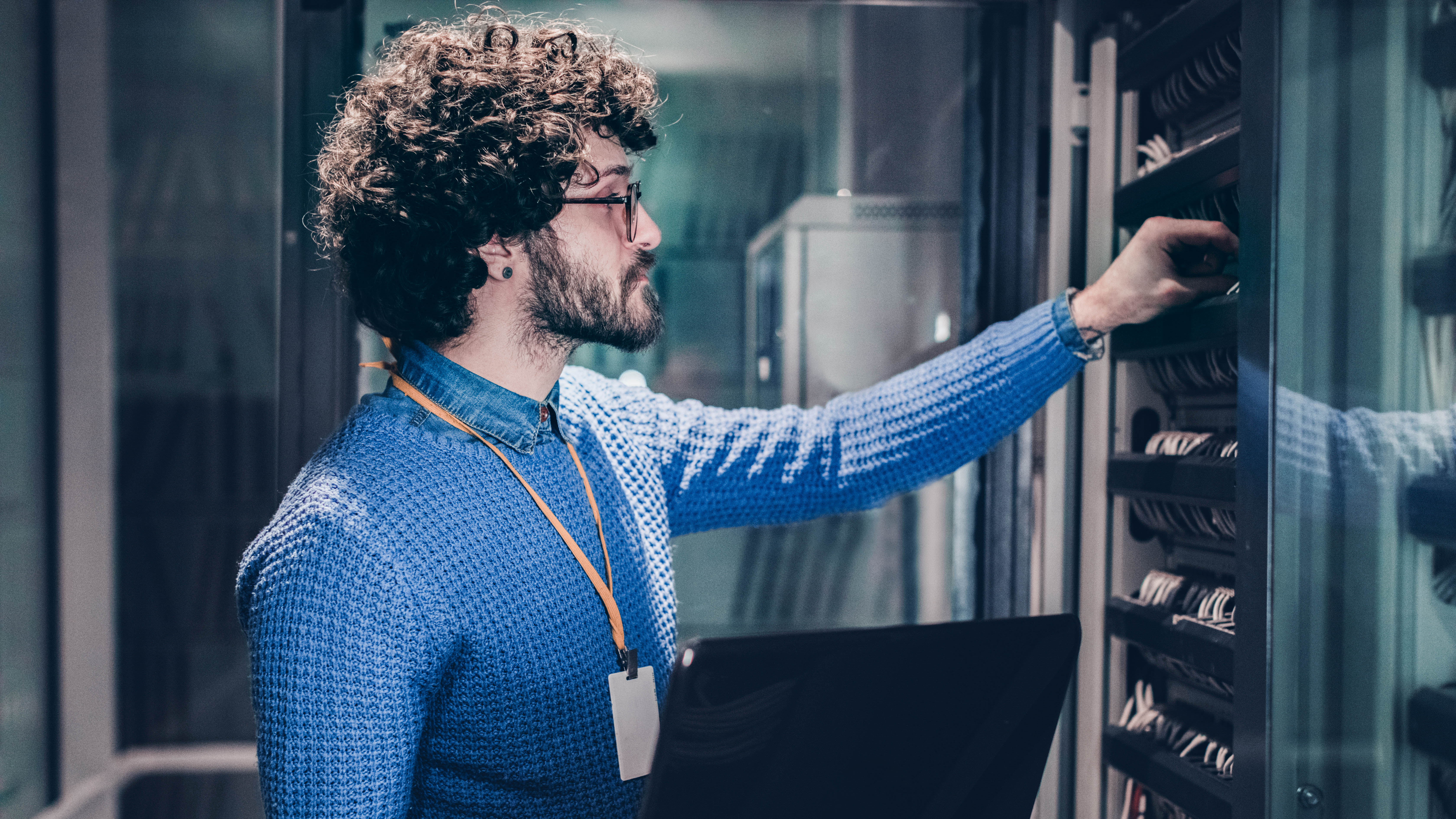 Mann mit lockigem Haar in blauem Pullover arbeitet an einem Serverrack in einem Rechenzentrum.