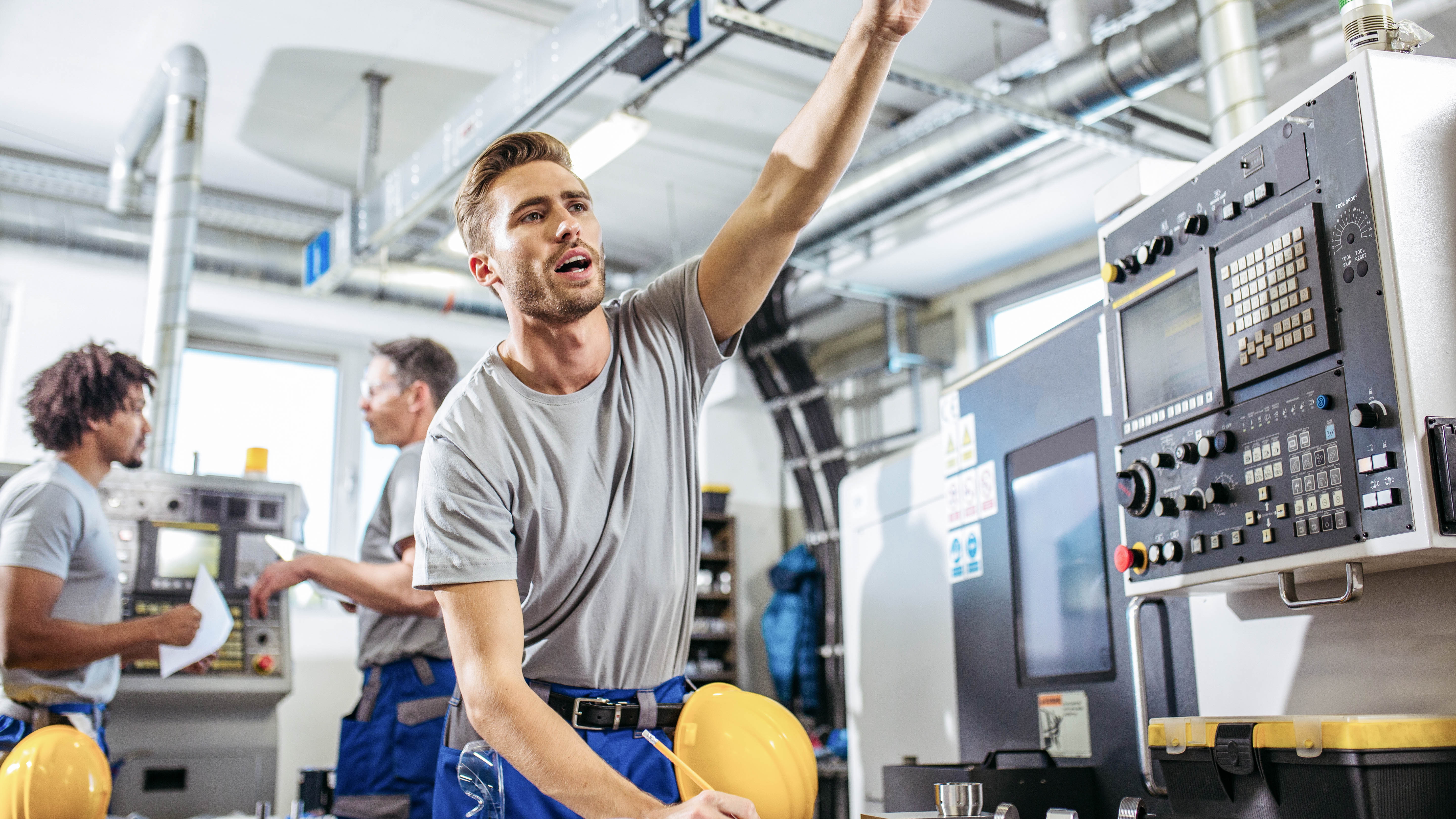 Ein Mann in Arbeitskleidung erklärt etwas, während zwei weitere Personen in einer Werkstatt an Maschinen arbeiten.