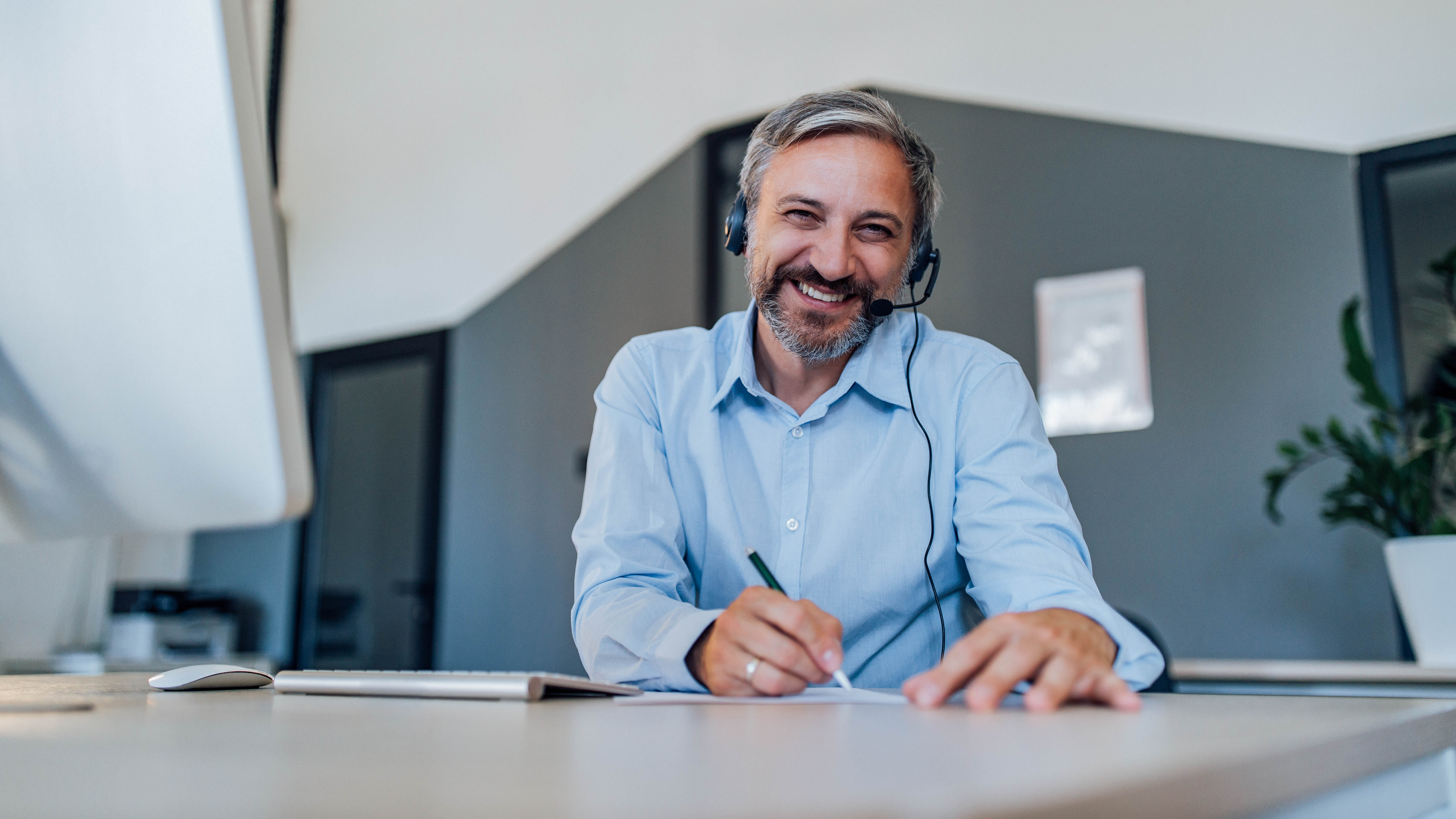 Mann mit Headset lächelt am Schreibtisch und schreibt mit einem Stift auf einem Blatt Papier.