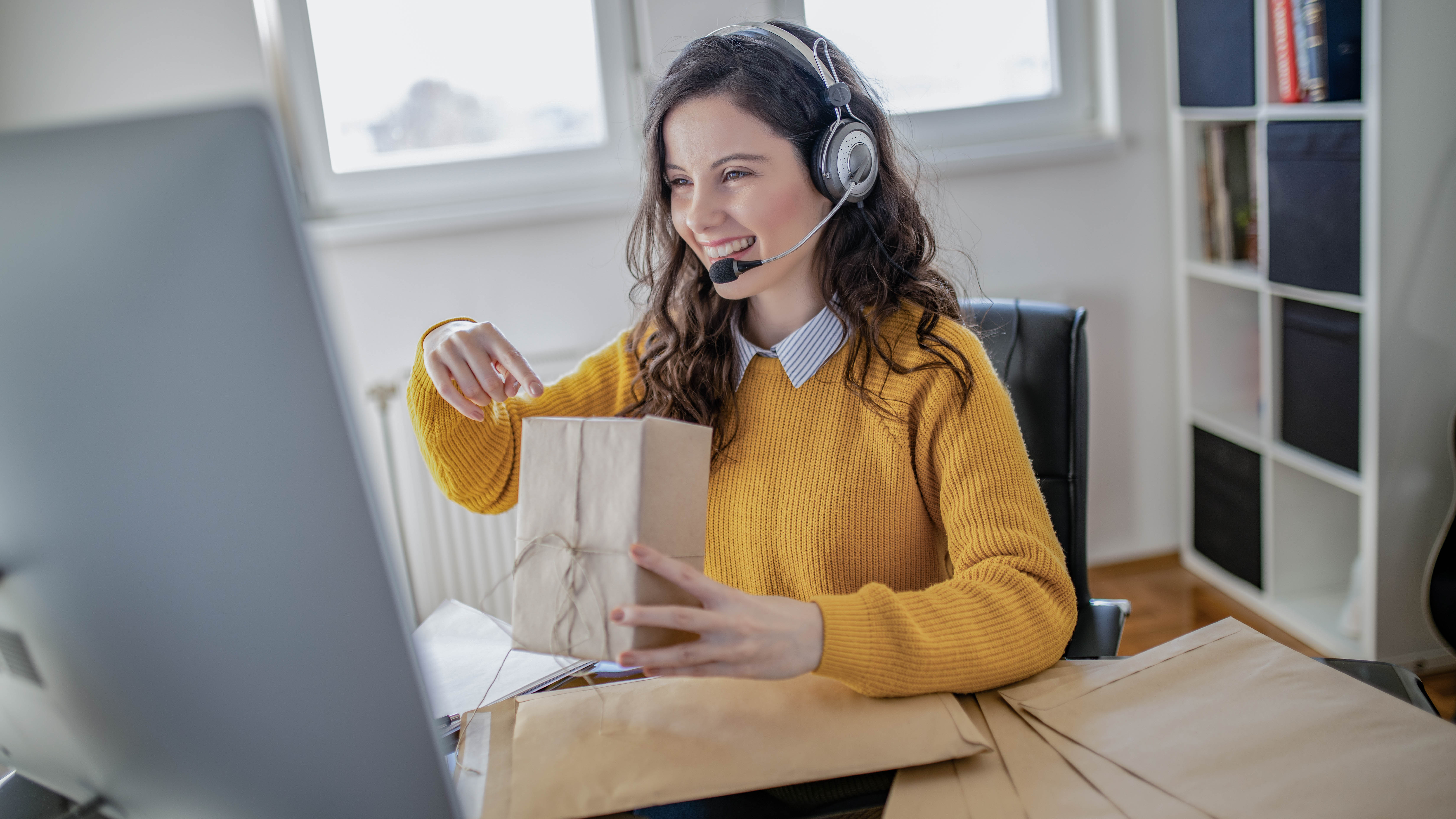 Frau mit Headset sitzt am Schreibtisch und hält ein Paket in der Hand, während sie auf den Computerbildschirm schaut.