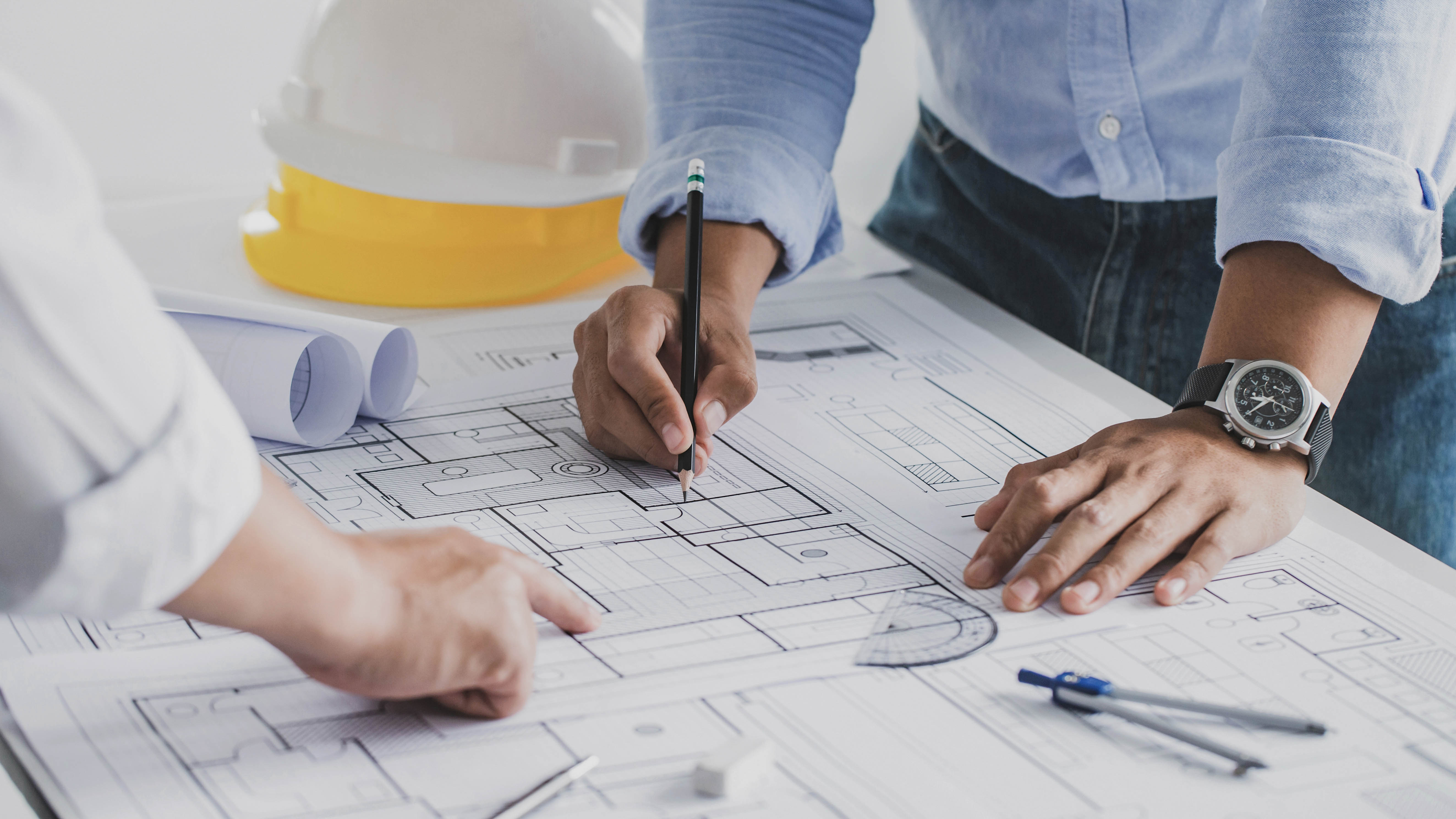 Hände von zwei Personen, die auf einem Bauplan arbeiten, mit einem Helm und Schreibutensilien im Hintergrund.