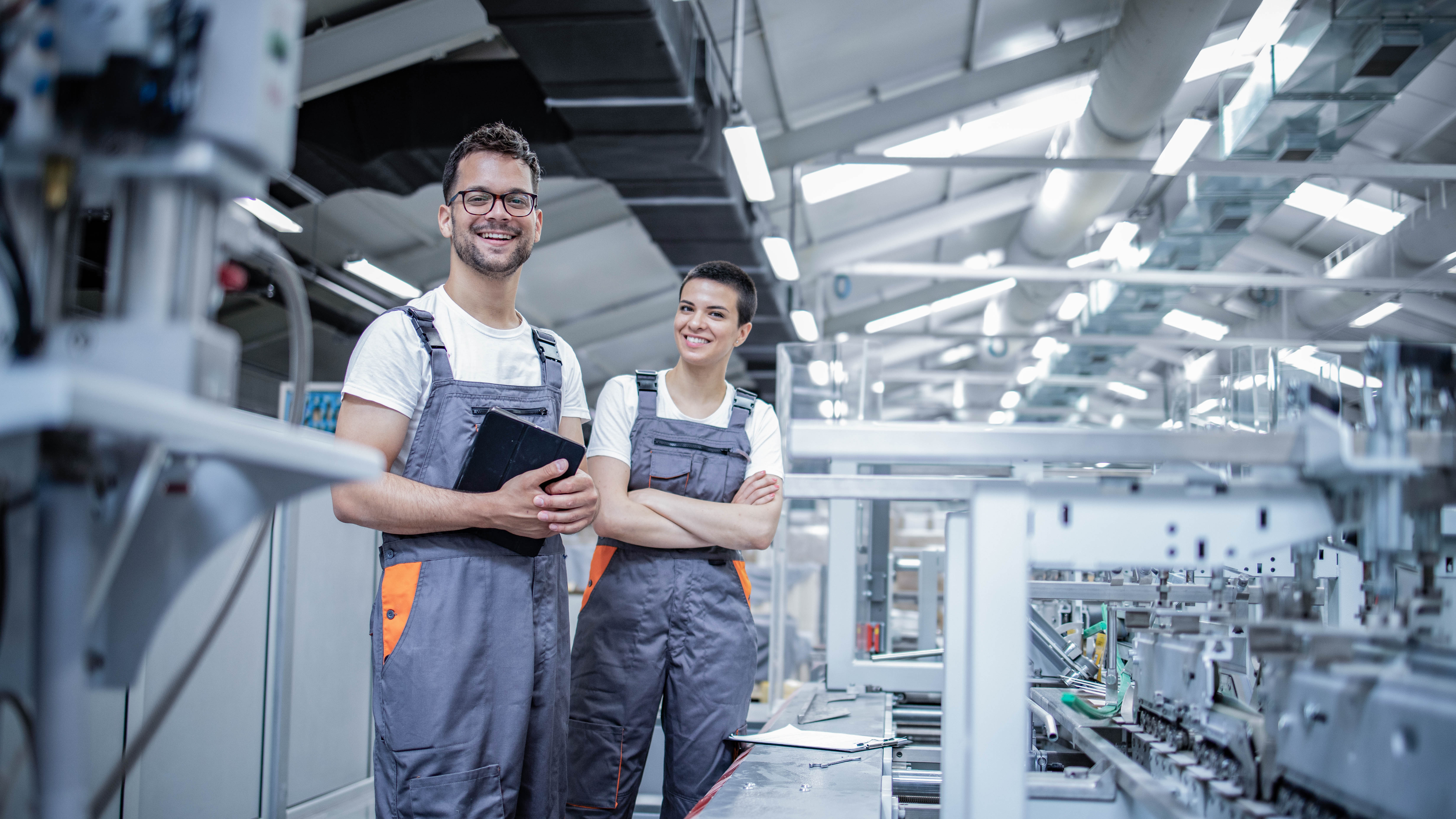 Zwei Personen in Arbeitskleidung stehen in einer modernen Produktionshalle und lächeln in die Kamera.
