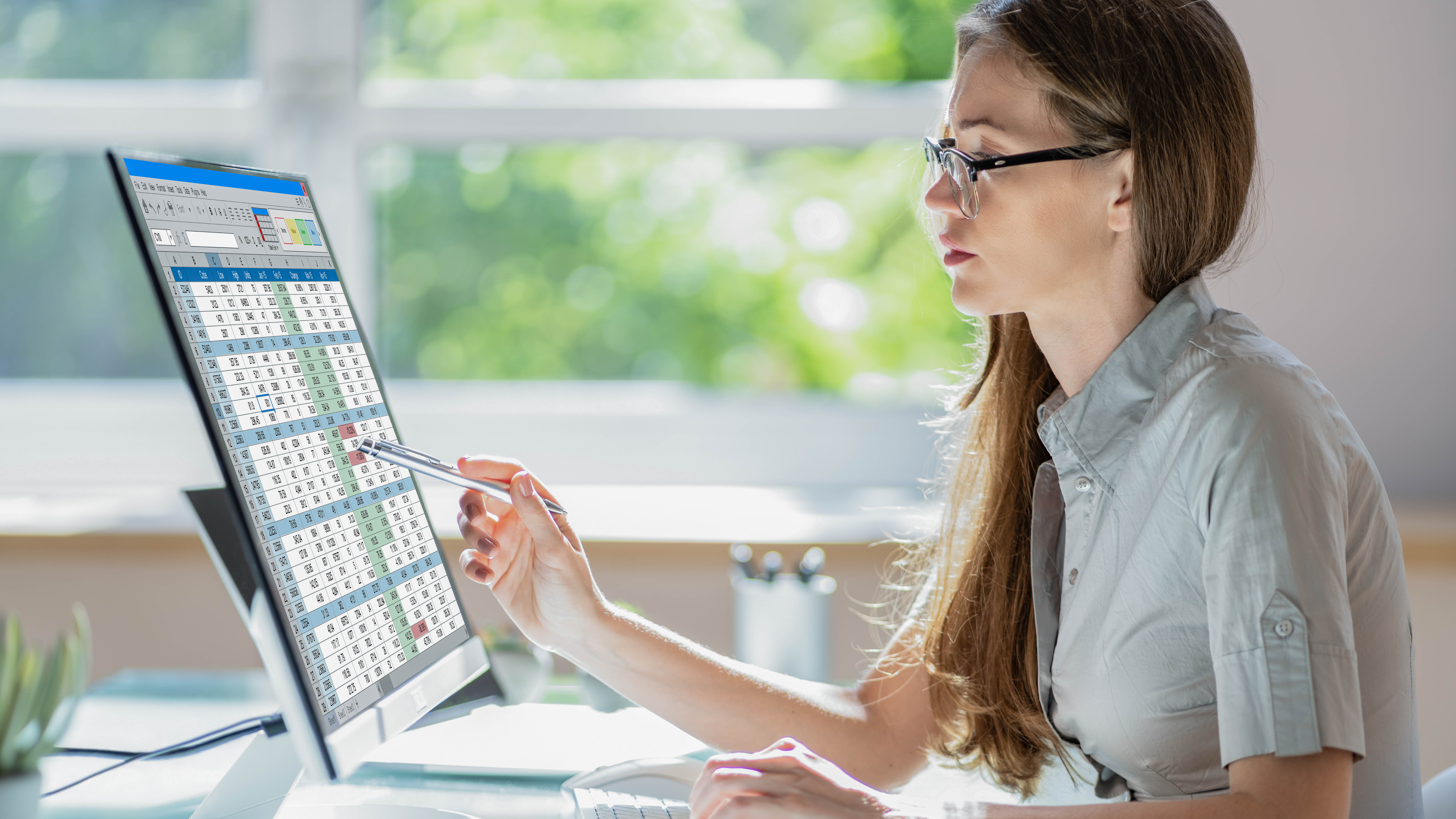 Eine Frau mit Brille betrachtet einen Computerbildschirm mit einer Tabelle und hält einen Stift in der Hand.