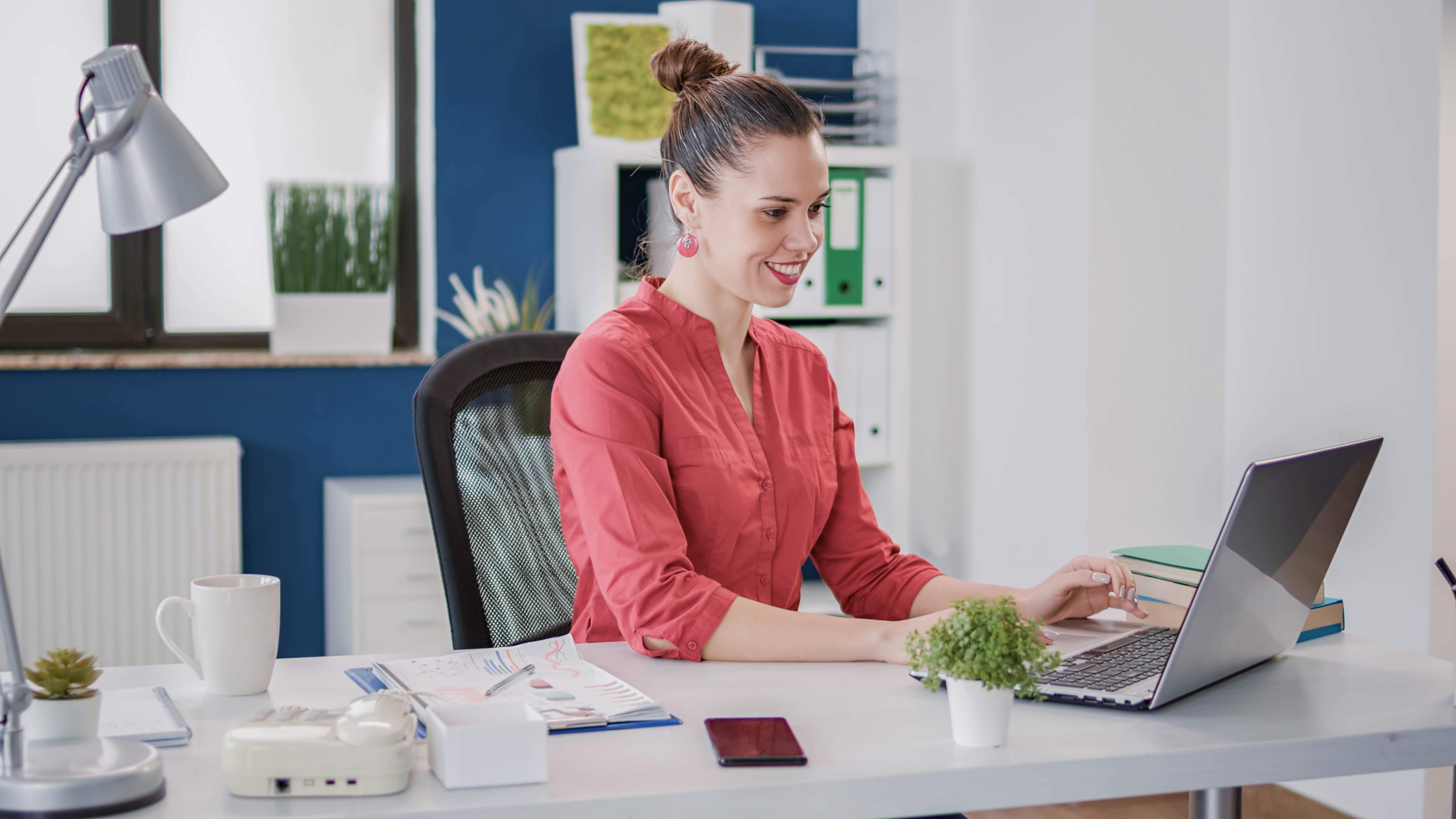 Eine Frau mit einem Dutt sitzt an einem Schreibtisch und arbeitet an einem Laptop, umgeben von Büromaterialien.