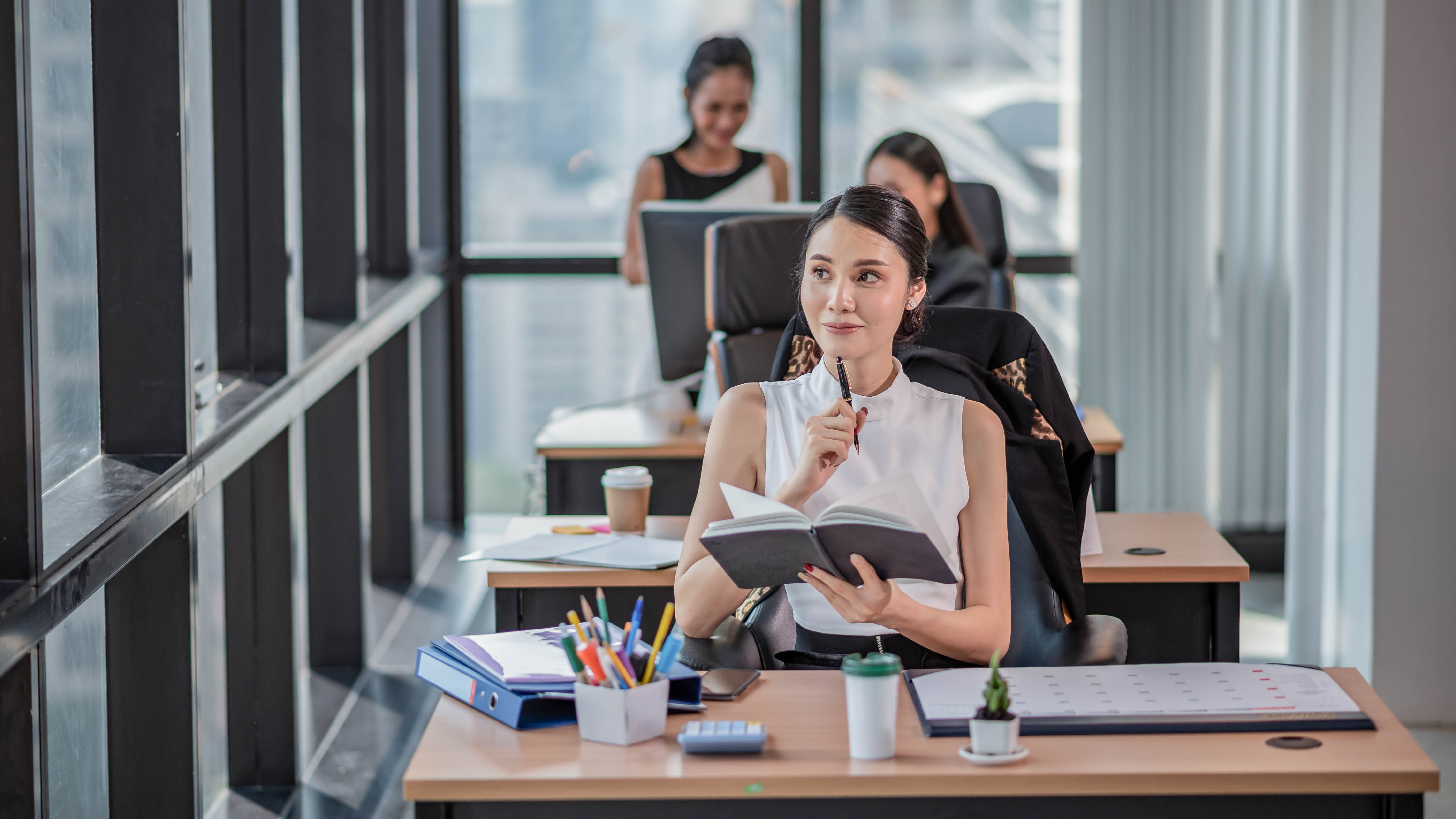 Eine Frau mit einem Notizbuch sitzt an einem Schreibtisch in einem modernen Büro, während andere Personen im Hintergrund arbeiten.