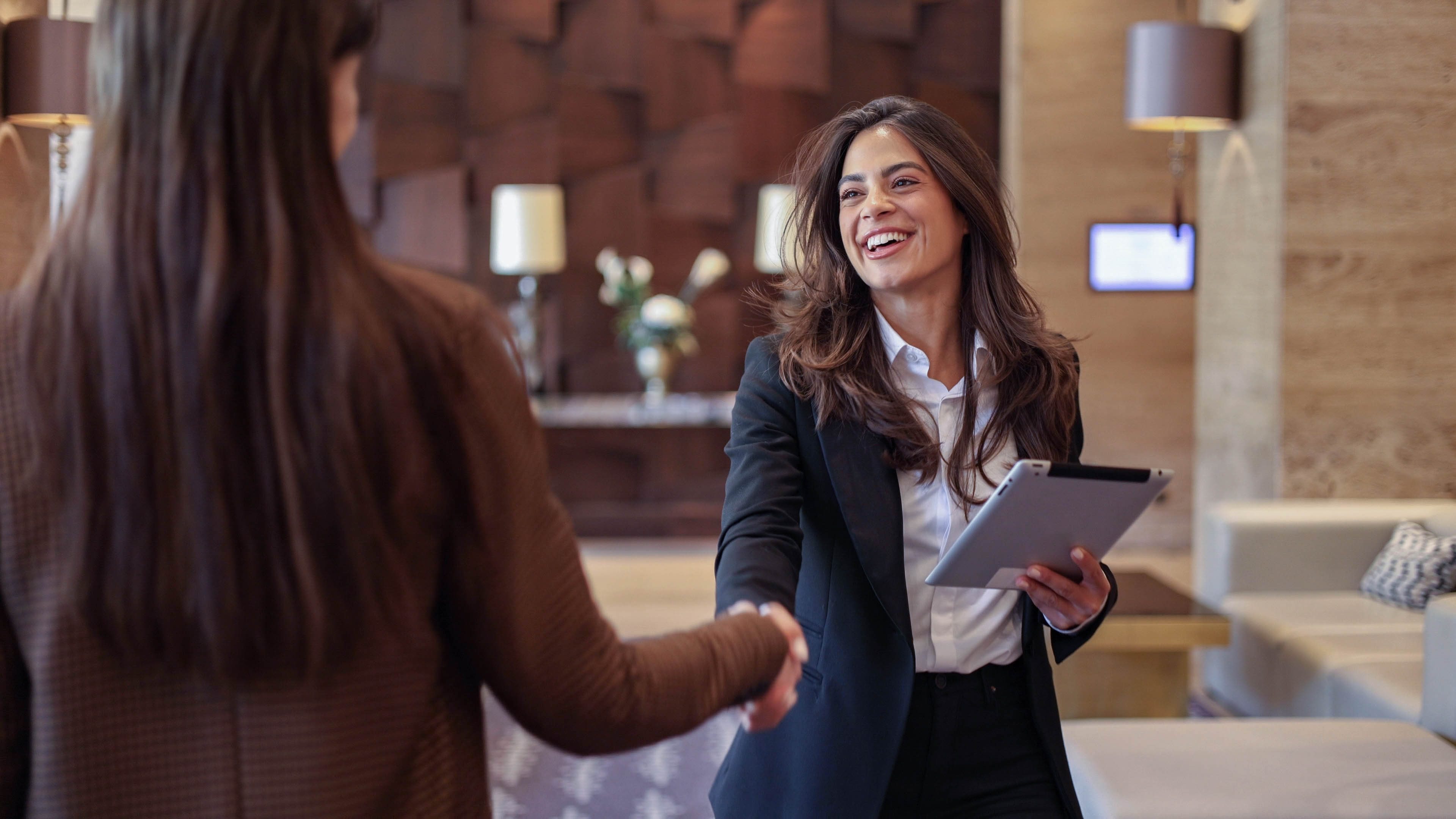 Zwei Frauen schütteln sich die Hände in einer modernen Lobby, eine hält ein Tablet.