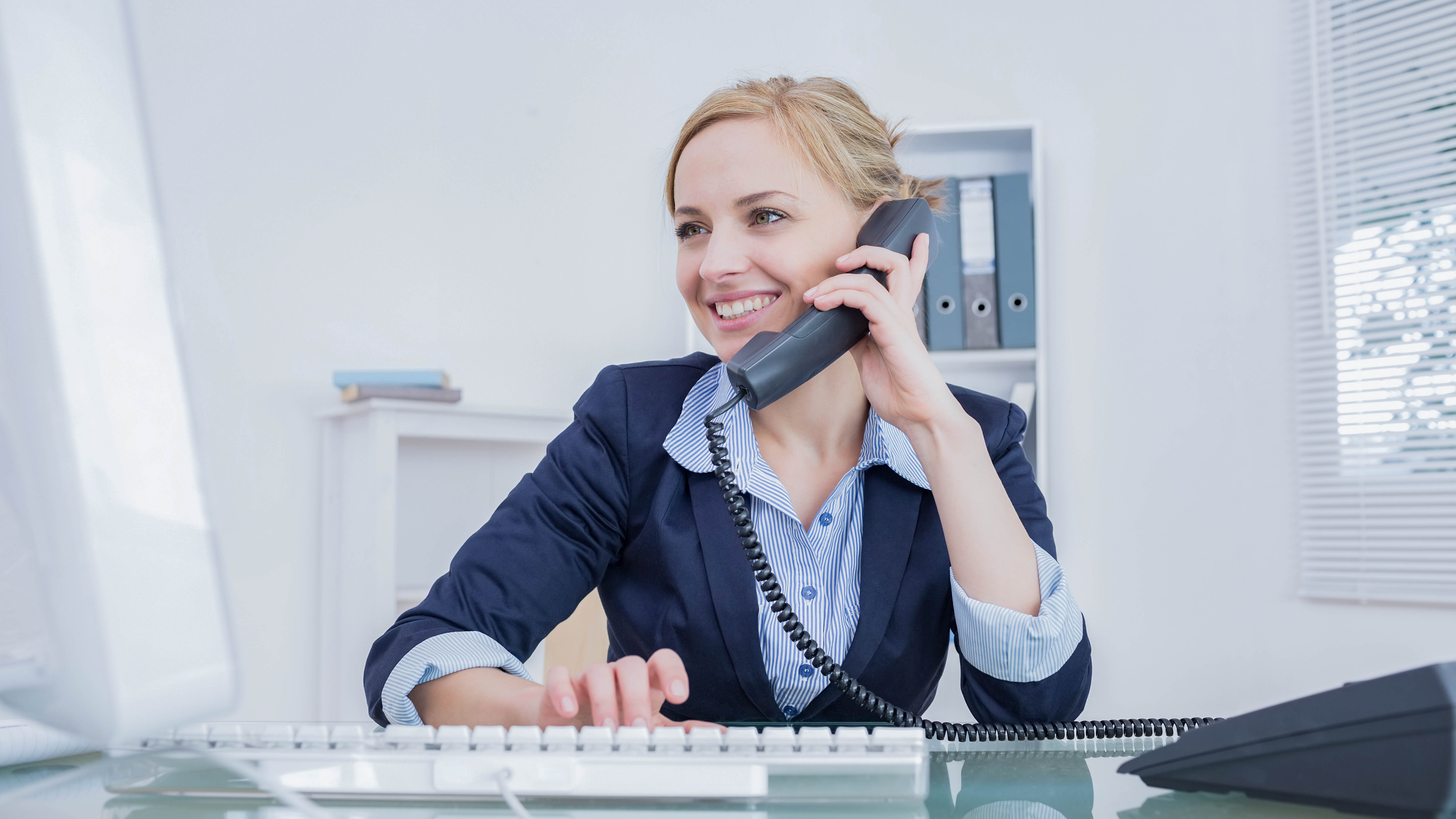 Frau mit Telefonhörer am Schreibtisch, lächelnd, während sie auf einer Tastatur tippt.