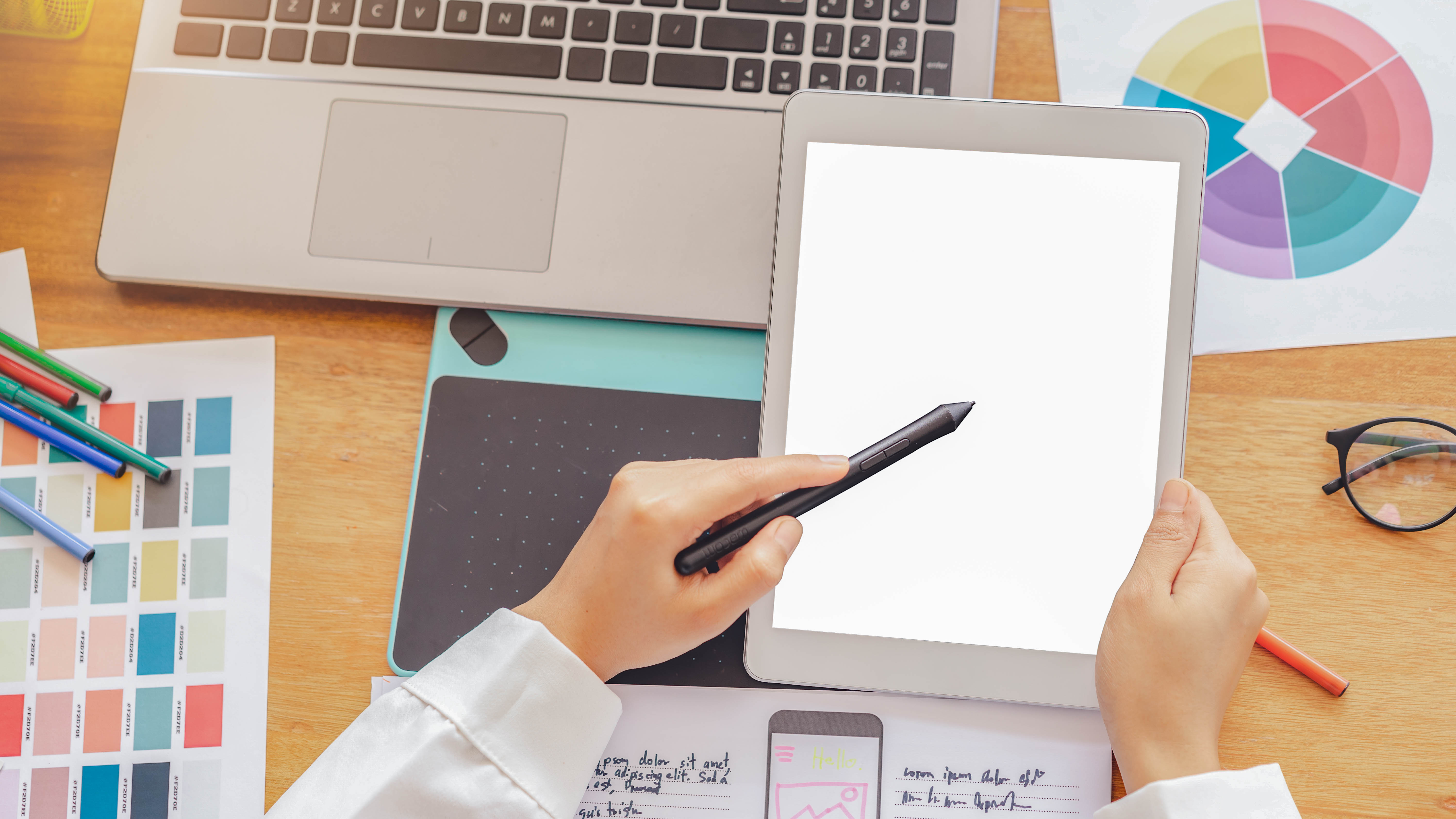 Eine Hand hält einen Stylus über ein leeres Tablet, umgeben von einem Laptop, Stiften und Farbmuster.