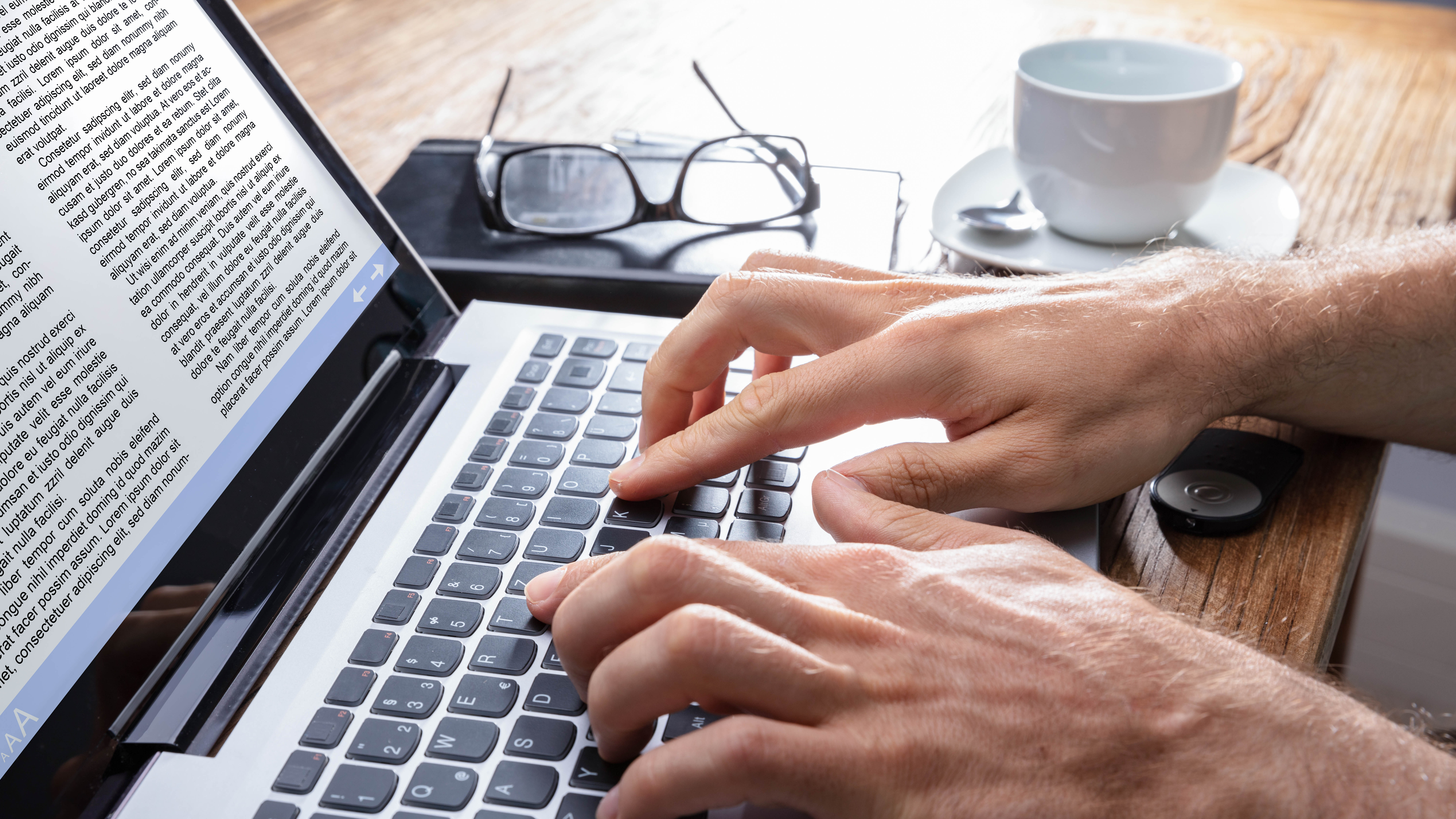 Hände tippen auf einer Laptop-Tastatur, im Hintergrund eine Tasse Kaffee und eine Brille.