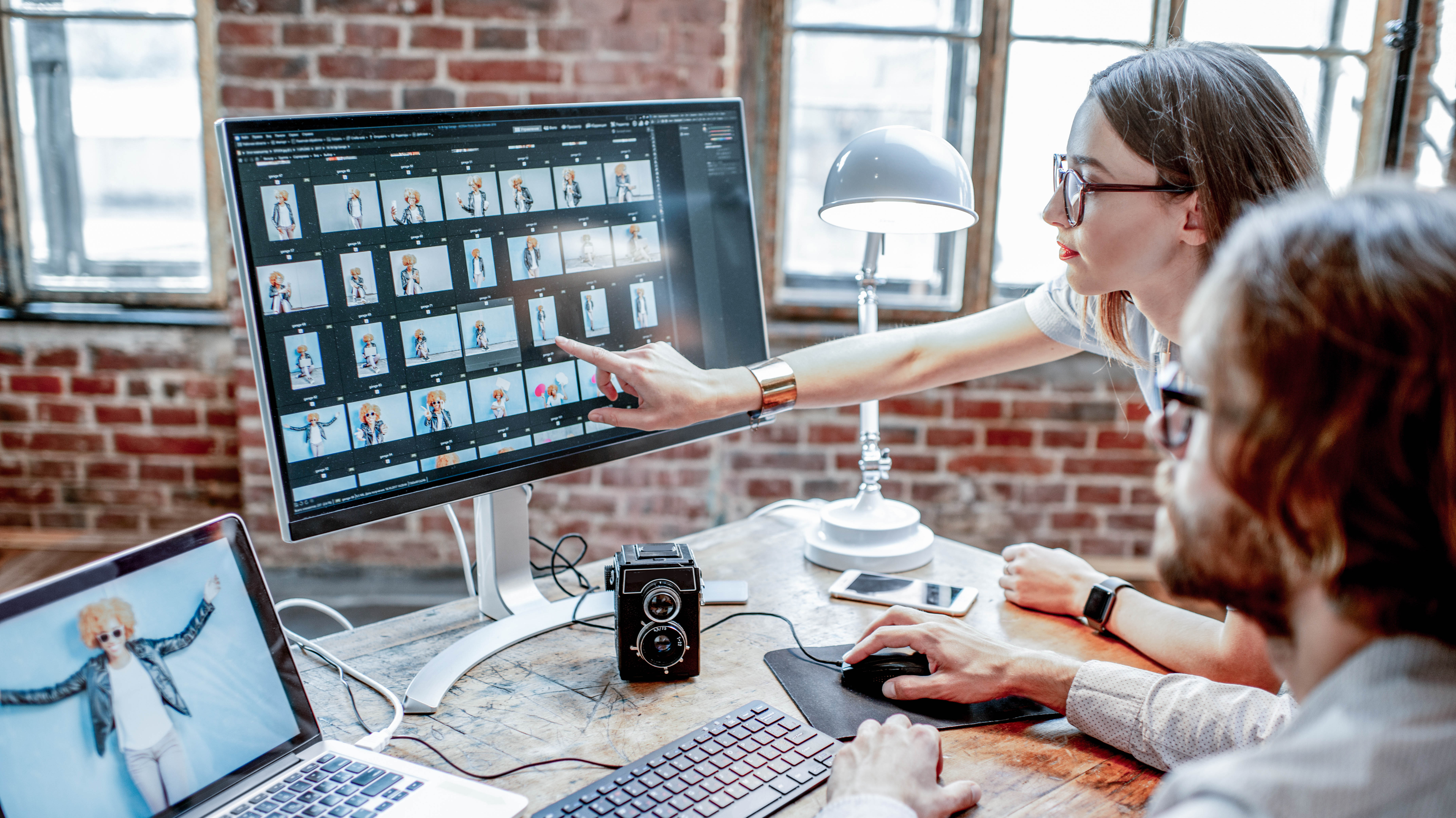 Eine Person zeigt auf einen Computerbildschirm mit Fotos, während eine andere Person am Tisch arbeitet.