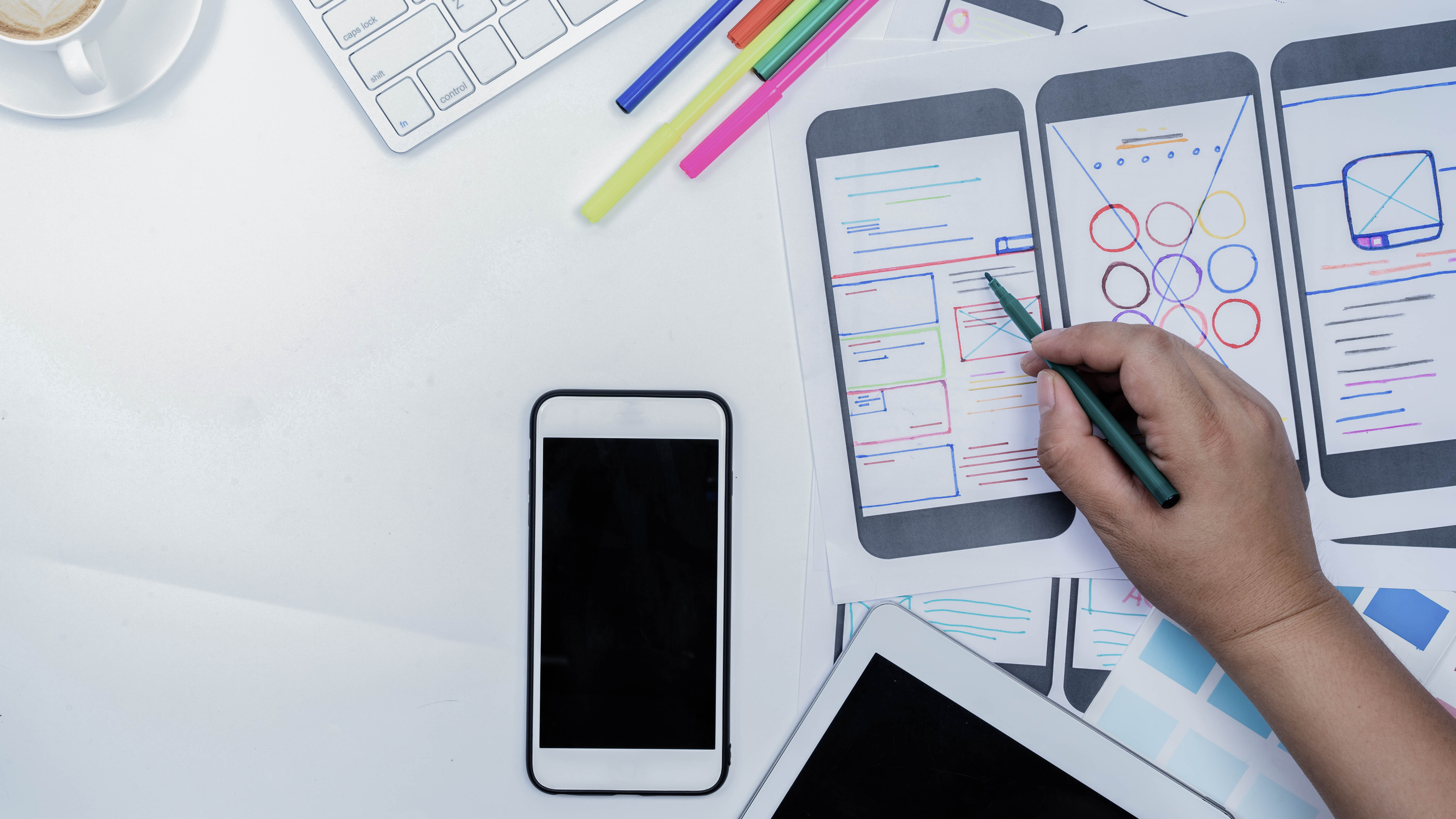 Auf einem Tisch liegen Skizzen von App-Layouts, ein Smartphone, ein Tablet, bunte Stifte und eine Tasse Kaffee.