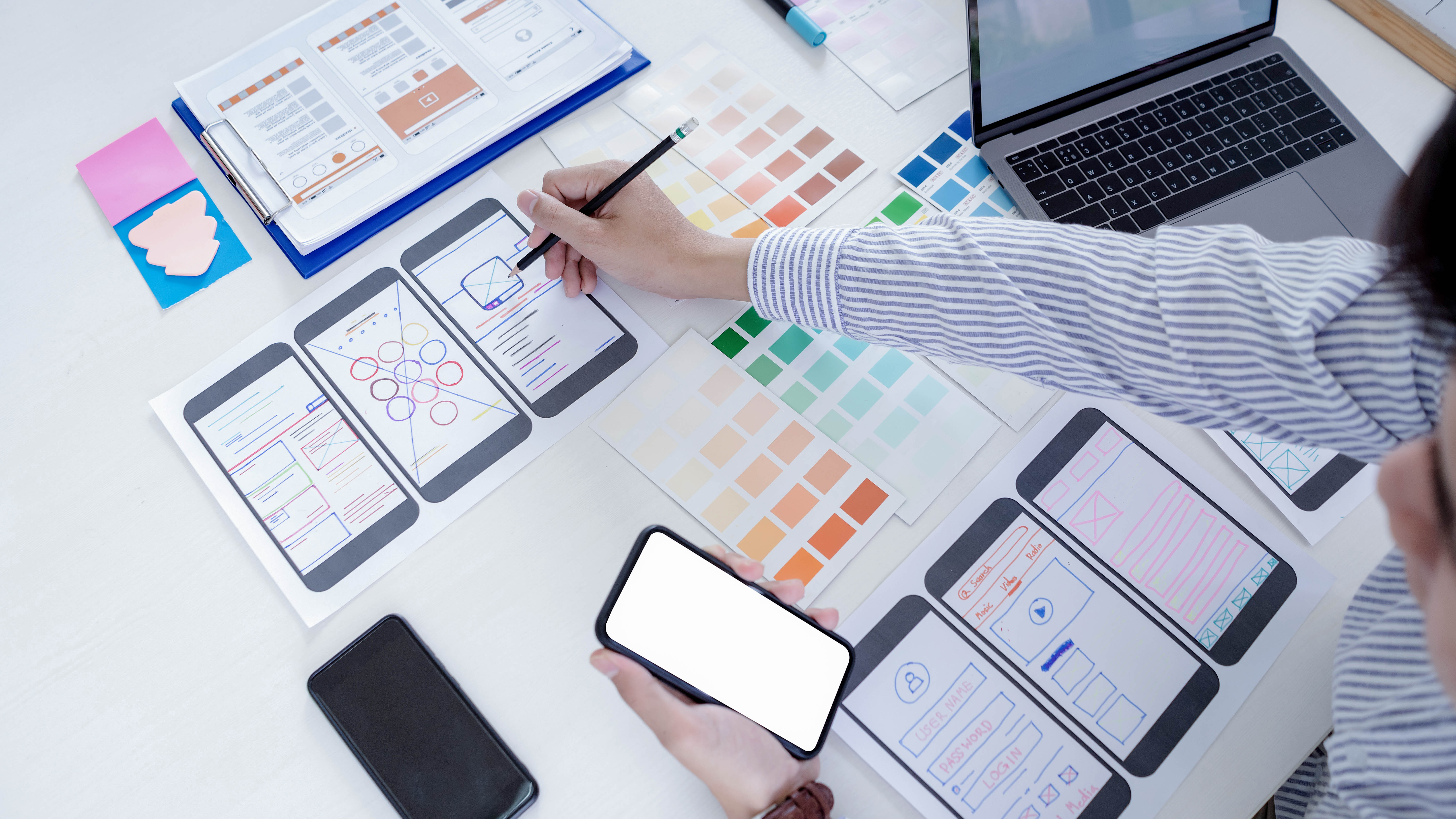Papierprototypen von Smartphone-Apps, Farbpaletten und ein Laptop auf einem Tisch, während eine Person daran arbeitet.
