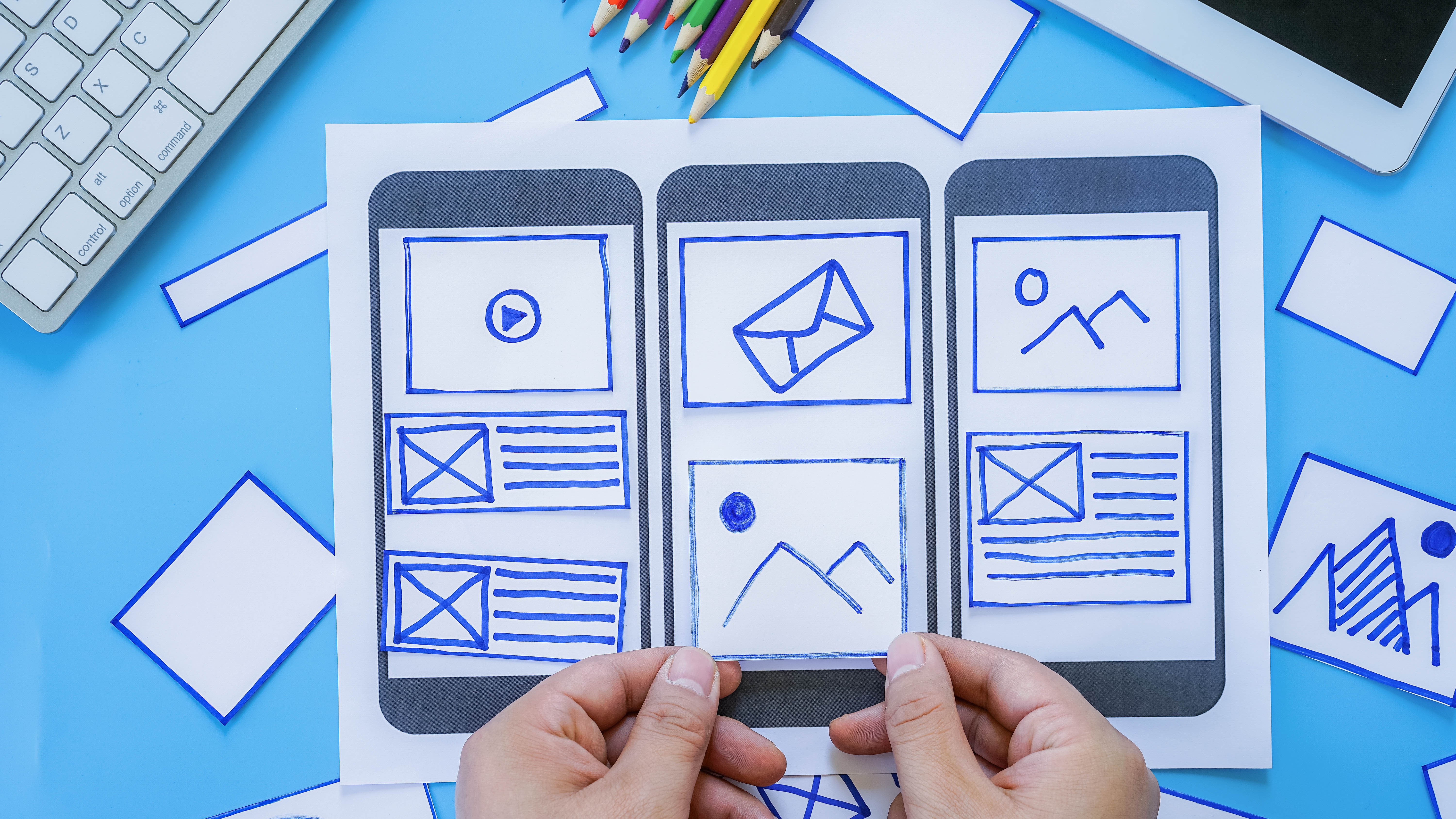 Hände halten ein Papier mit skizzierten Smartphone-Layouts, umgeben von bunten Stiften und weiteren Papierstücken.