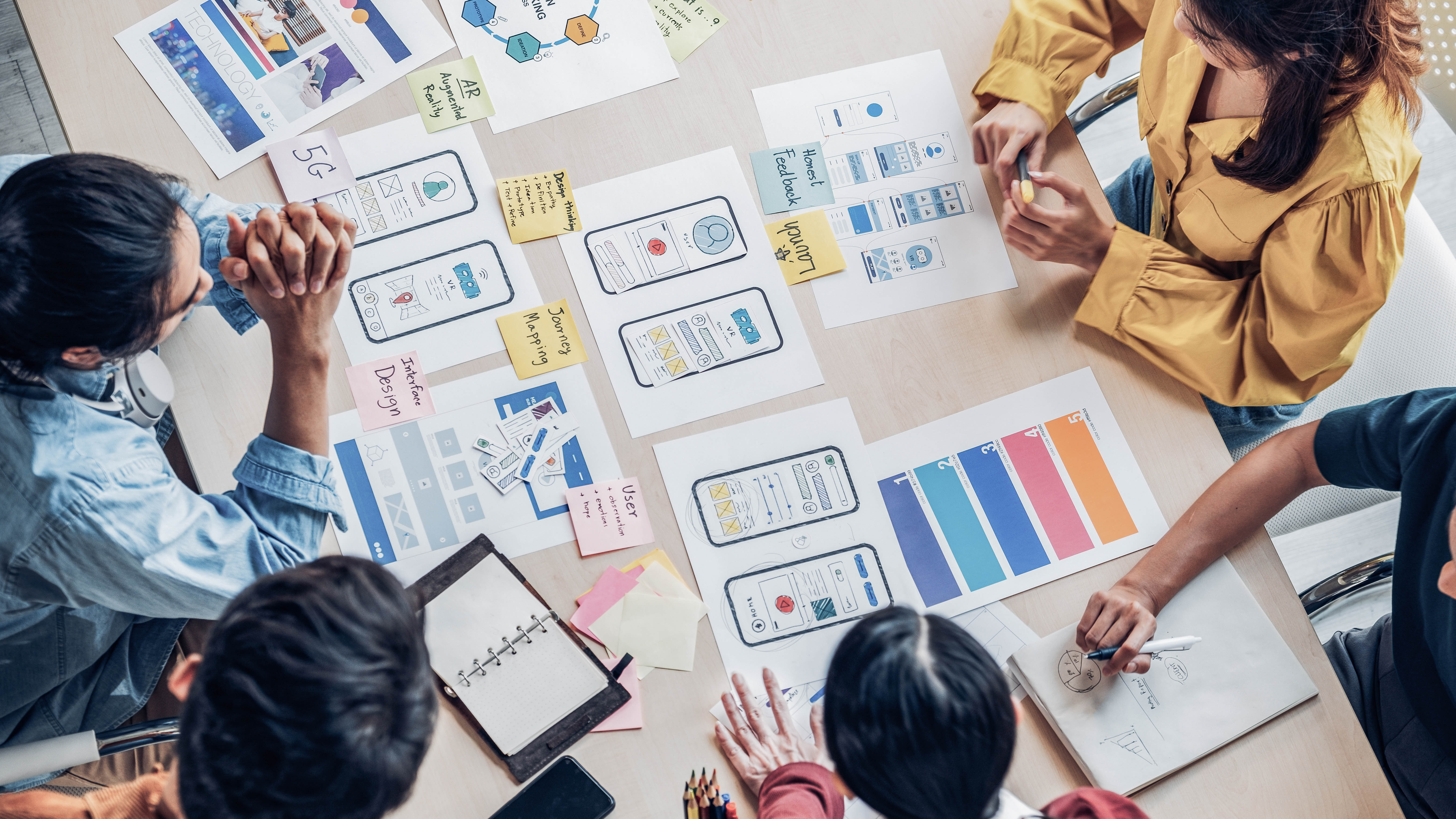 Mehrere Personen arbeiten an einem Tisch mit Skizzen von App-Designs, Notizen und Stiften.