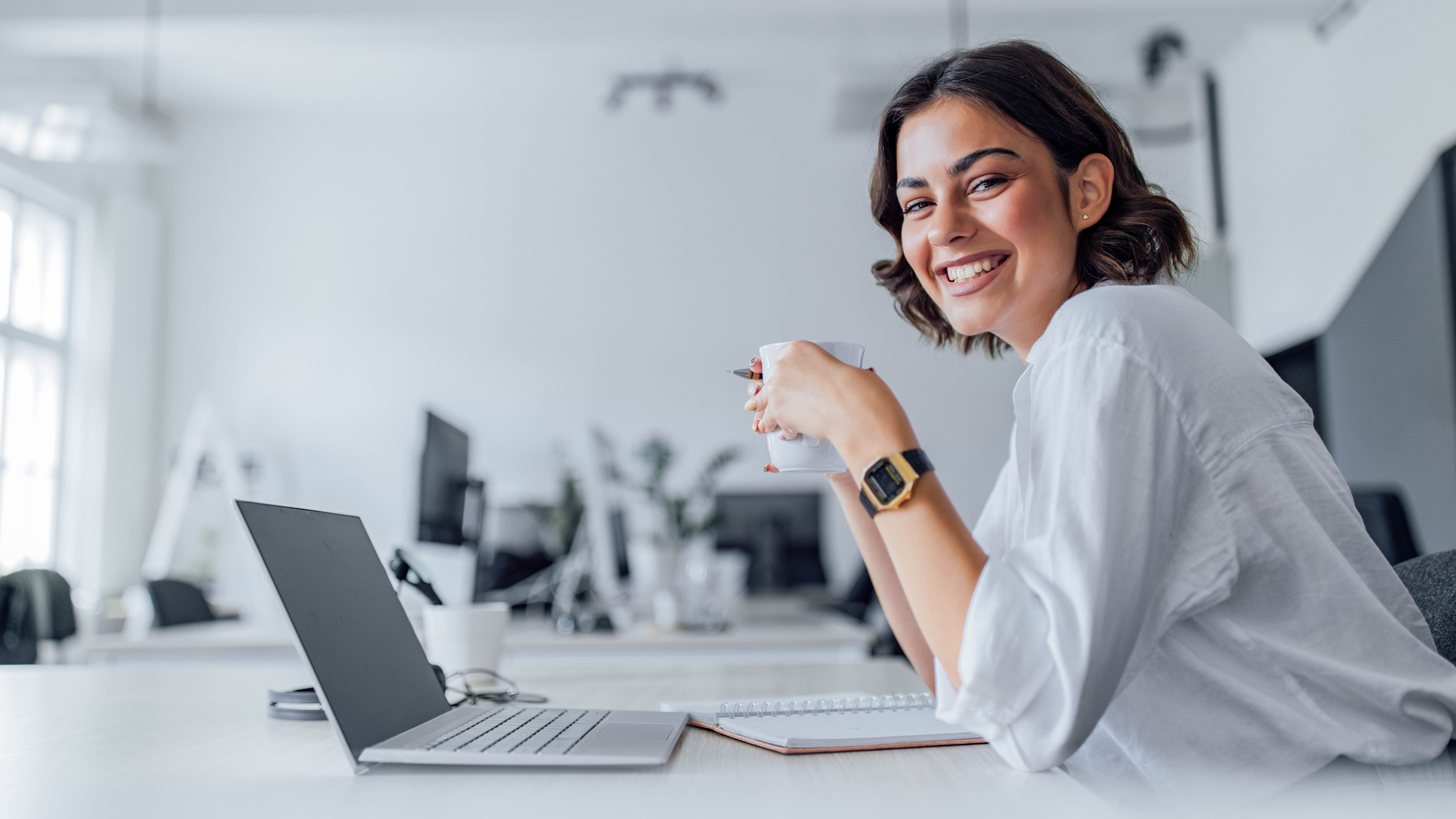 Junge Frau mit Kaffeetasse lächelt am Schreibtisch, Laptop und Notizbuch sind sichtbar.