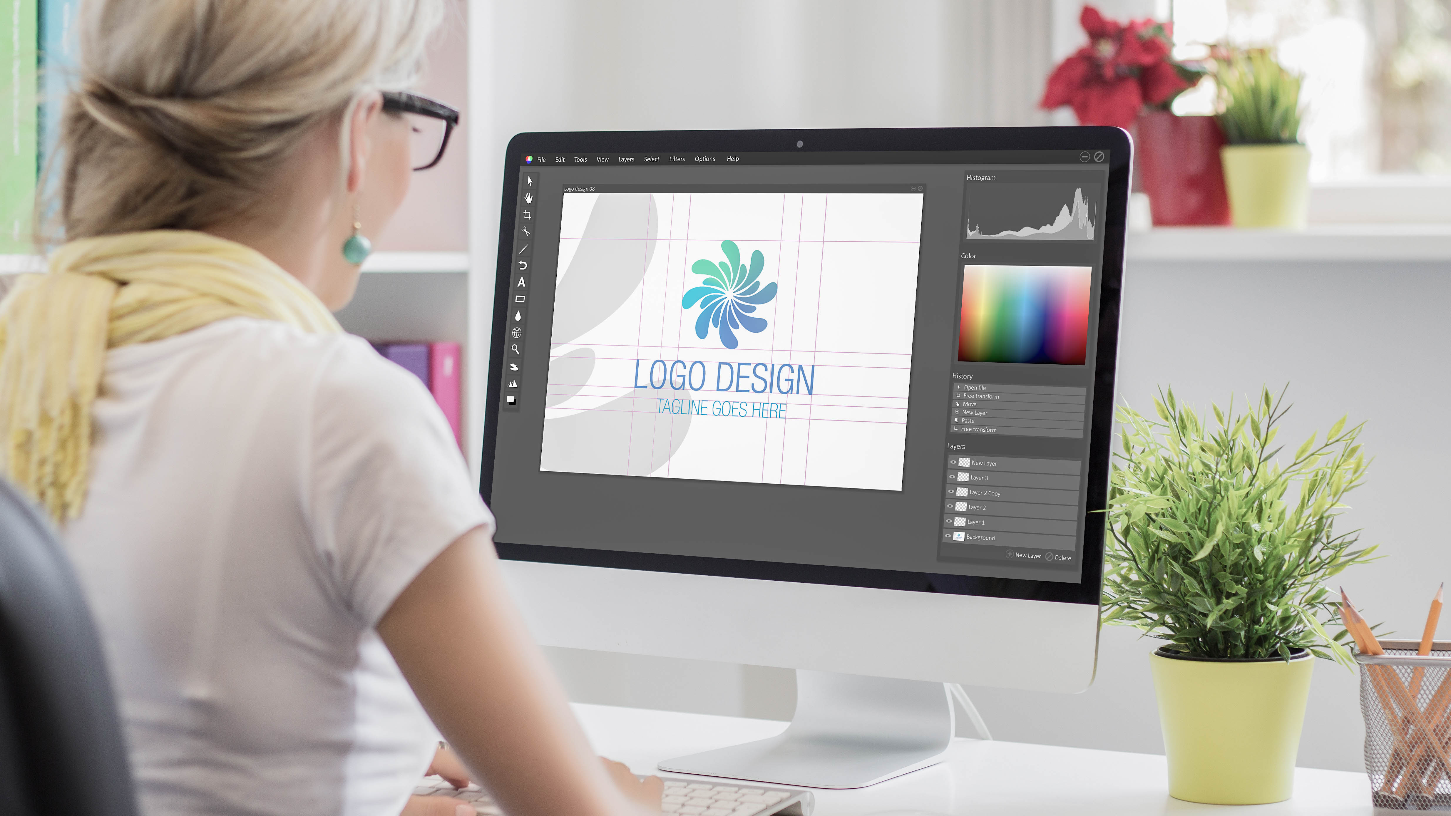 Eine Person arbeitet am Computer an einem Logo-Design mit verschiedenen Farben und einem grünen Pflanzentopf im Hintergrund.