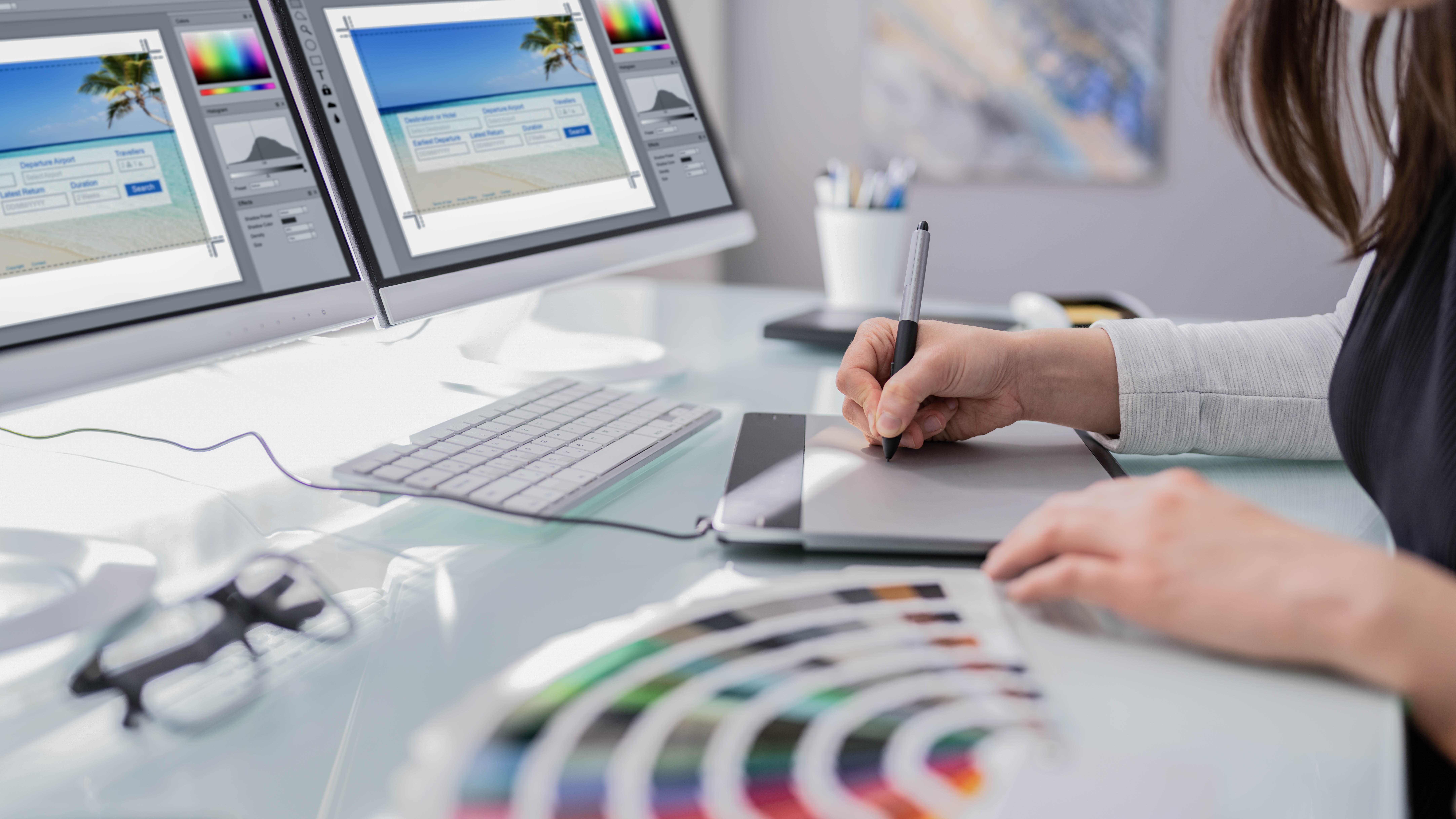 Eine Person zeichnet mit einem Grafiktablett, während zwei Monitore mit Grafiksoftware und Farbmustern im Vordergrund zu sehen sind.