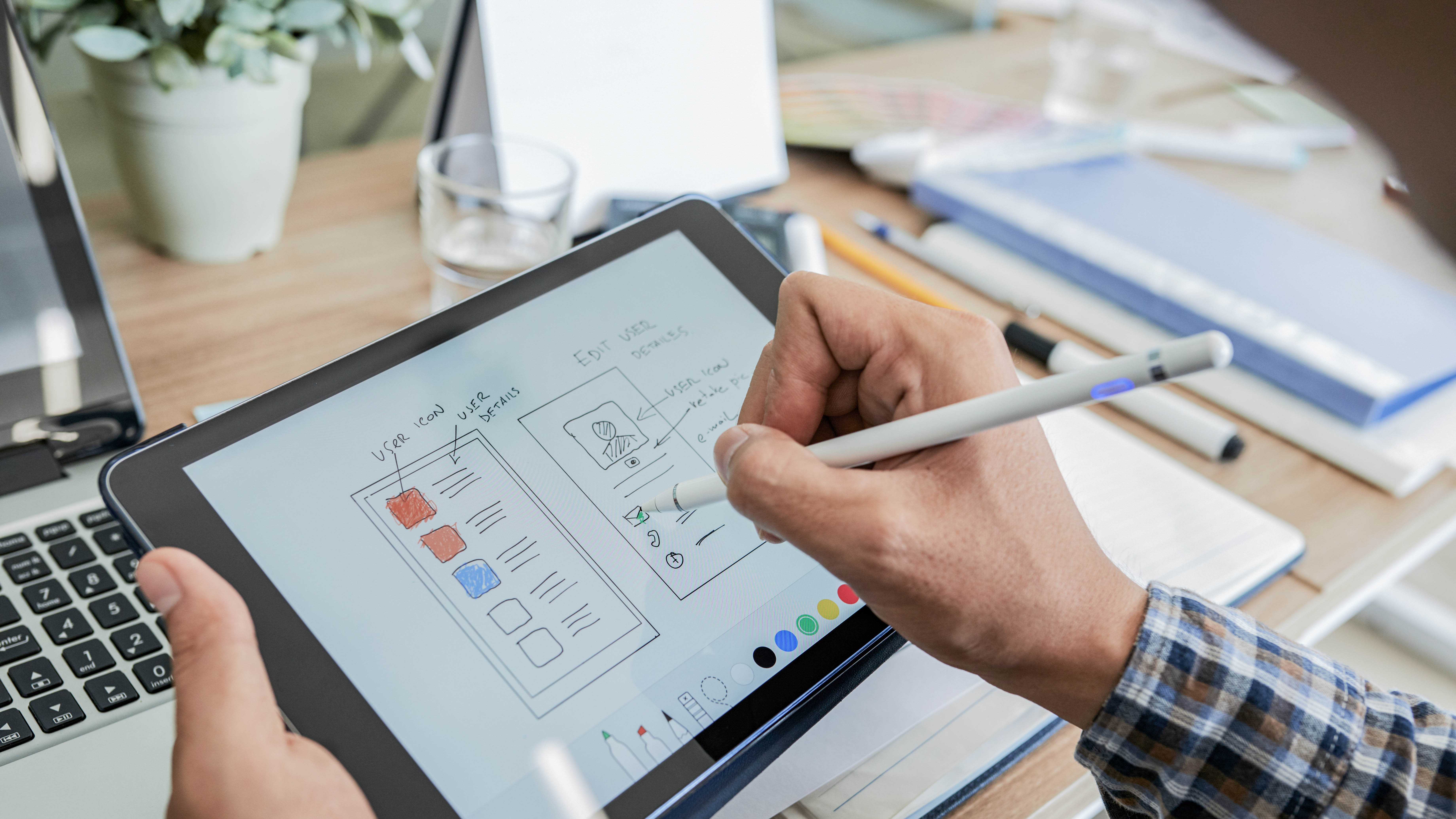Hand, die mit einem Stylus auf einem Tablet skizziert, um ein Benutzeroberflächendesign zu erstellen, im Hintergrund Bürobedarf.