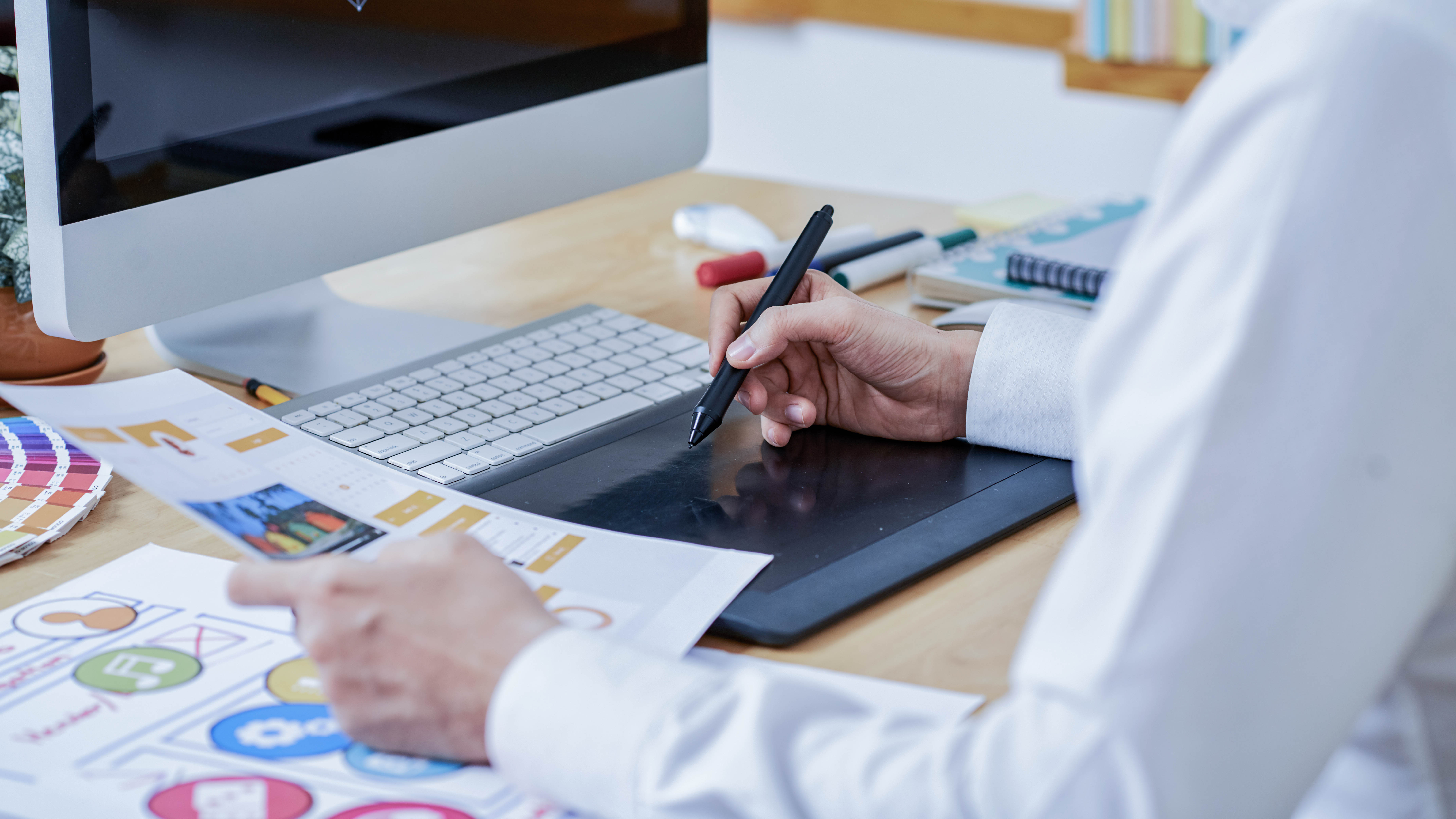 Eine Person hält einen Stift und arbeitet an einem Grafiktablett, während sie Unterlagen auf einem Tisch betrachtet.