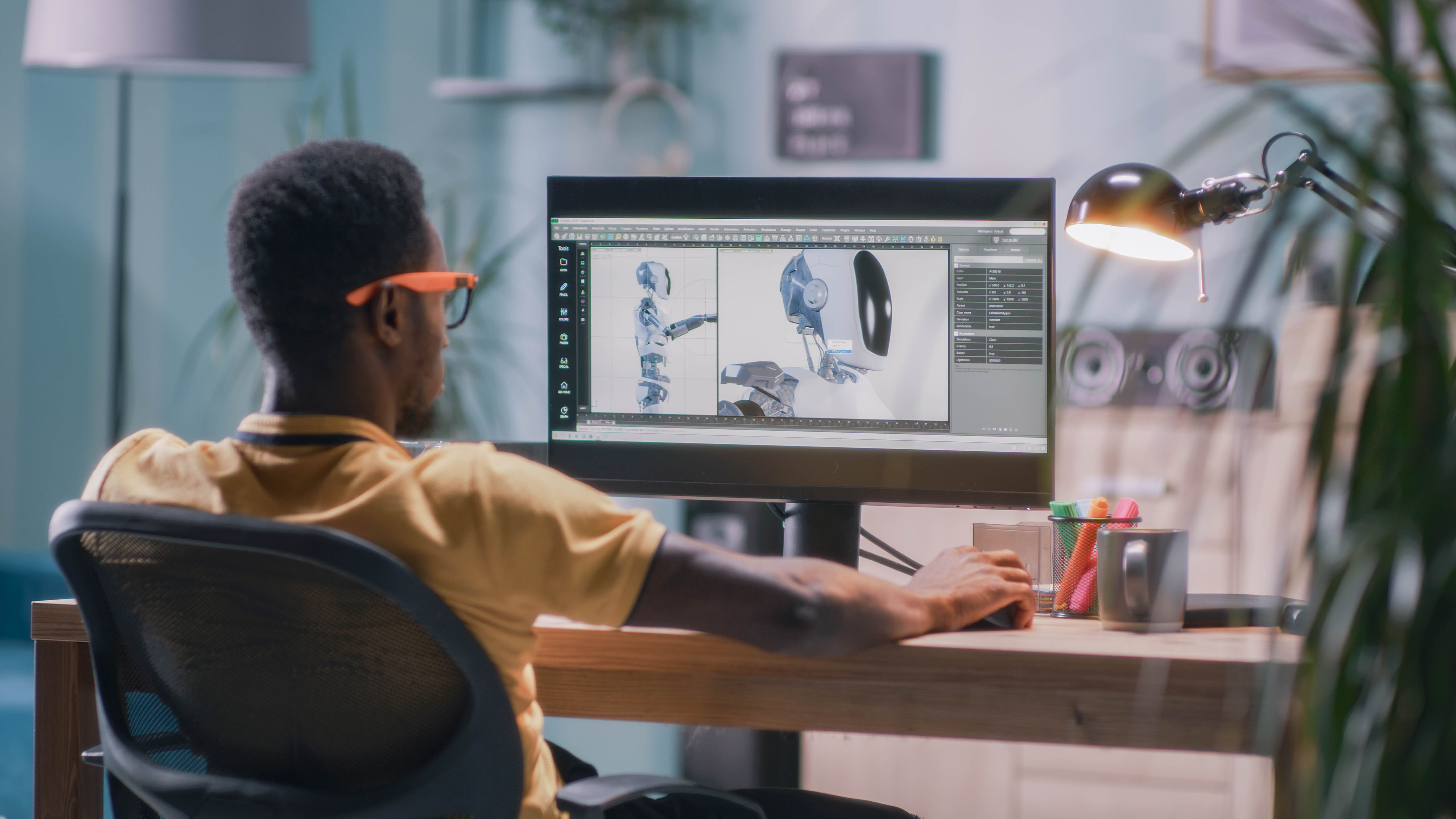 Ein Mensch mit Brille und orangefarbenem Rahmen sitzt an einem Schreibtisch und arbeitet an einem Computer mit 3D-Modellen.