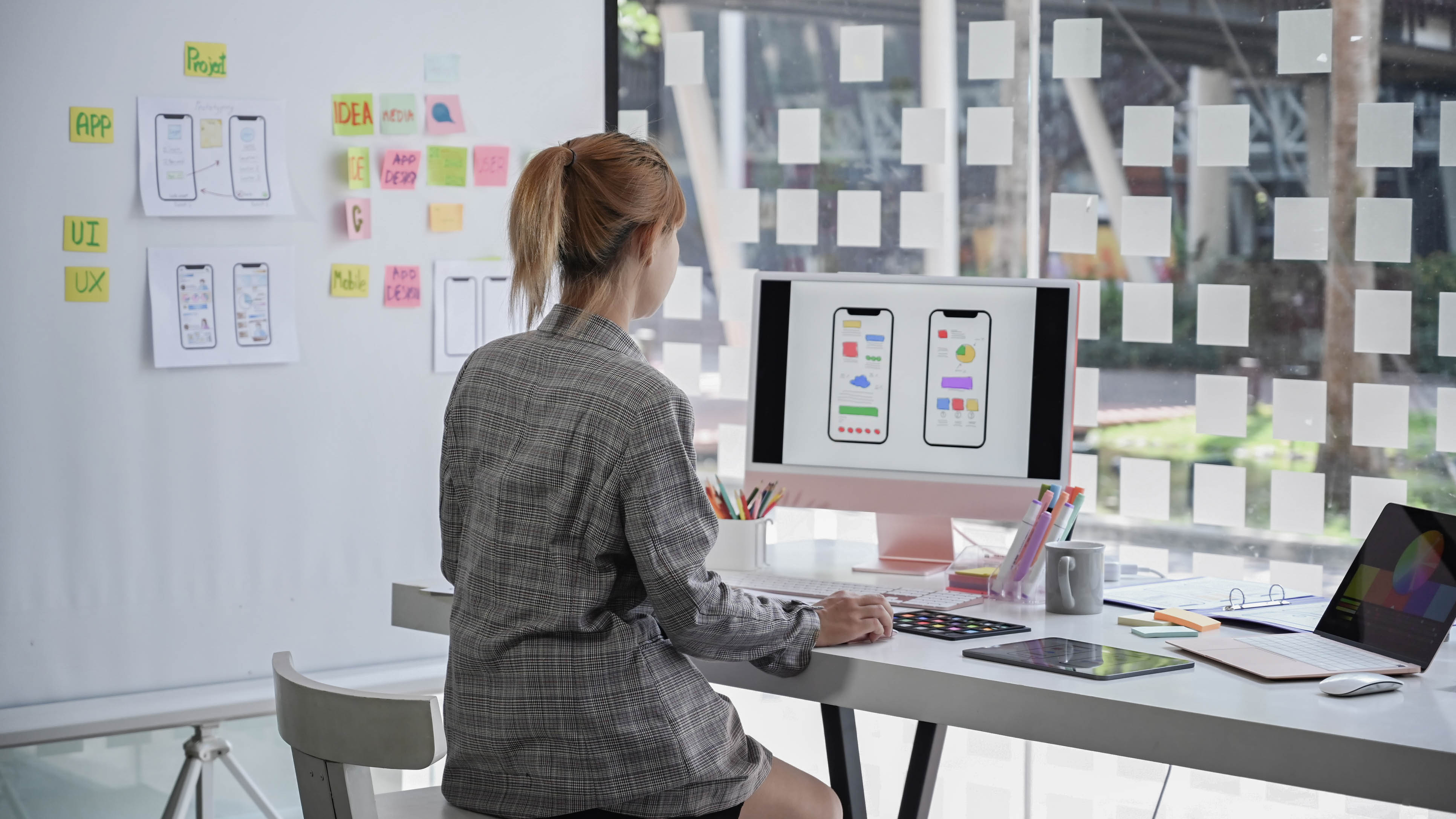 Eine Person sitzt an einem Schreibtisch und betrachtet zwei Smartphone-Displays mit App-Designs.