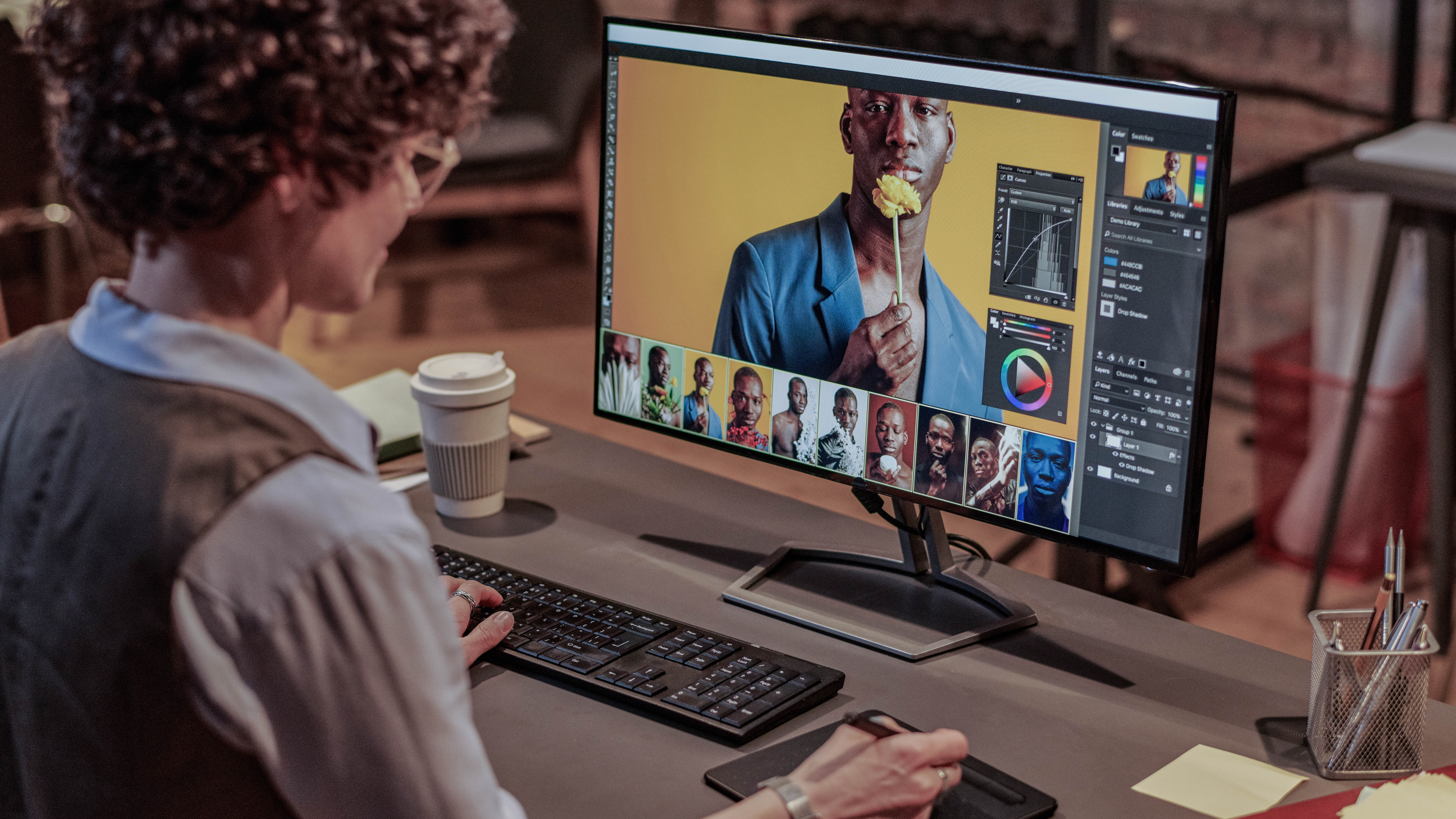 Eine Person mit lockigem Haar bearbeitet Bilder am Computer, auf dem ein Porträt eines Mannes mit einer Blume zu sehen ist.