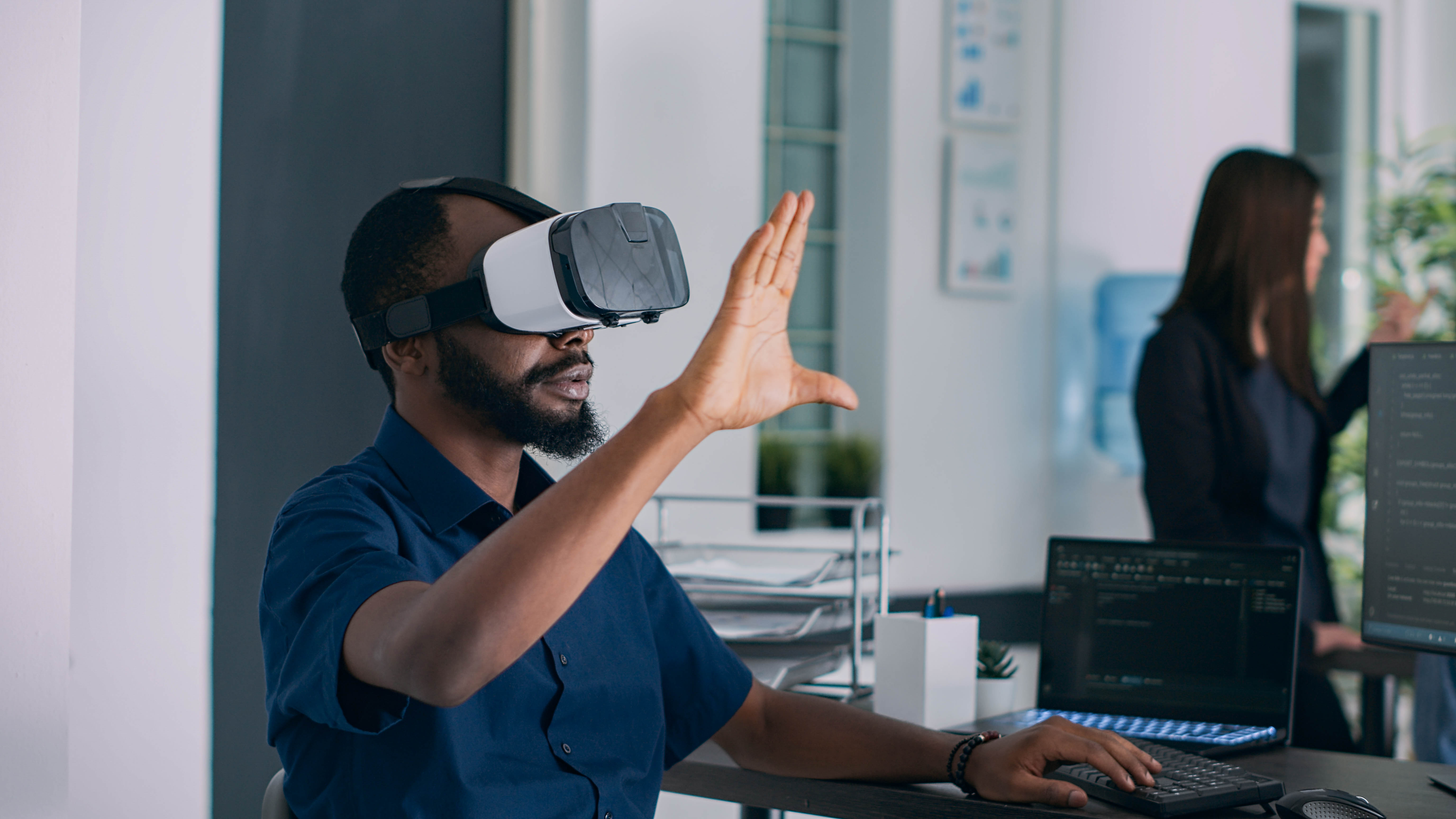 Mann mit VR-Brille hebt die Hand, während er an einem Schreibtisch mit Laptops und Büromaterial arbeitet.