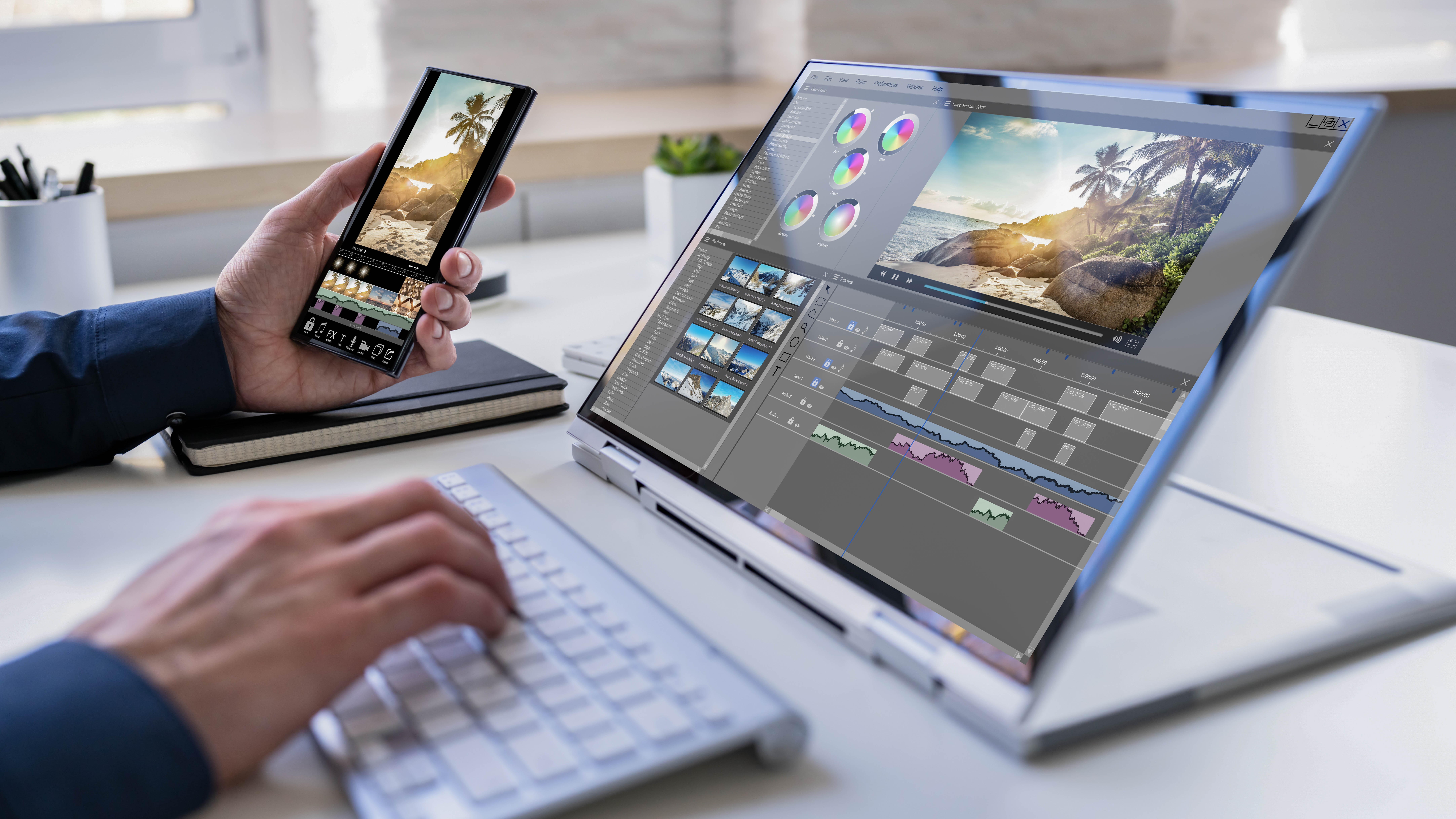 Eine Hand hält ein Smartphone, während die andere auf einer Laptop-Tastatur tippt; auf dem Bildschirm ist eine Bildbearbeitungssoftware sichtbar.