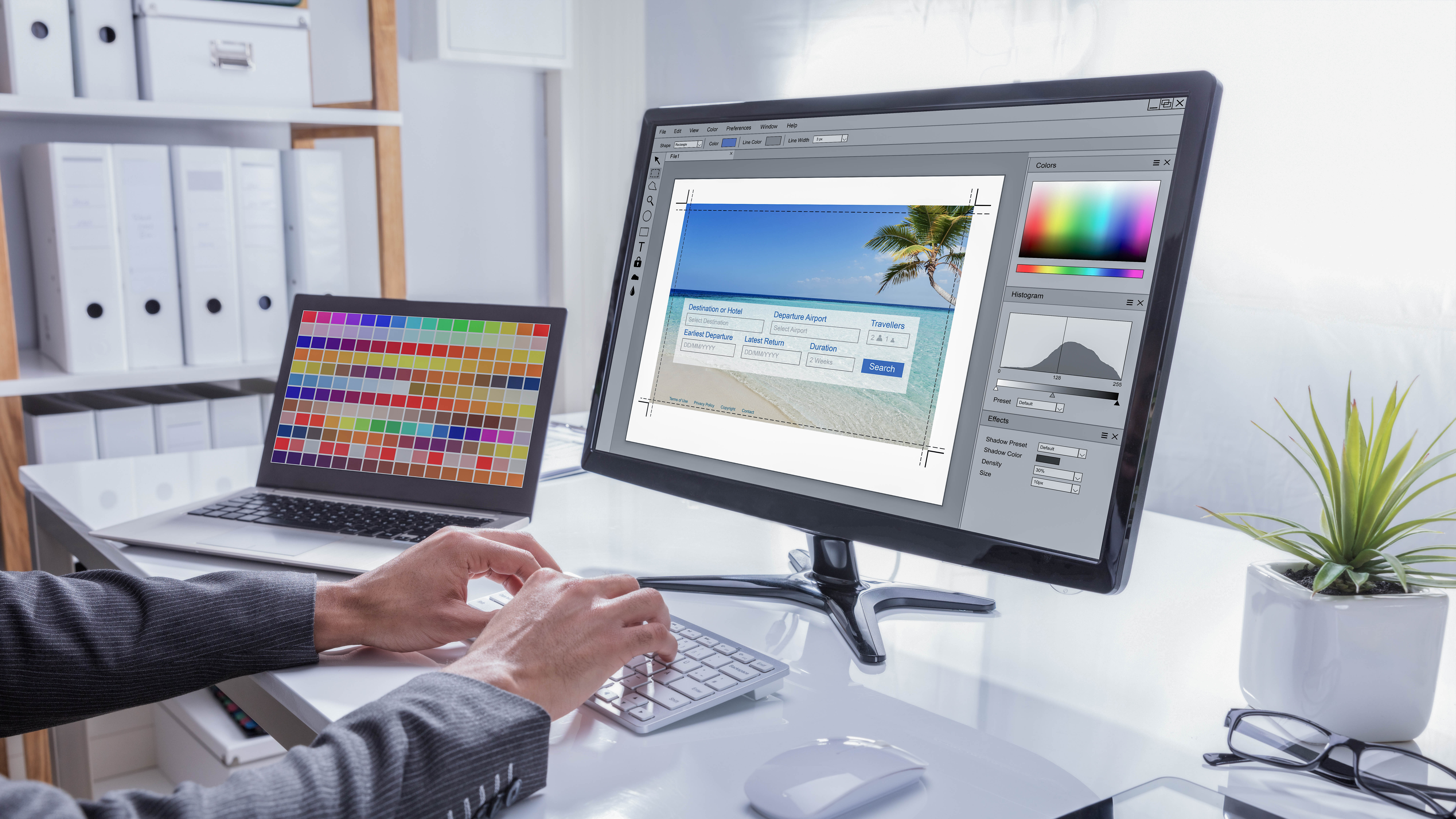 Ein Mensch arbeitet an einem Computer mit einem Grafikprogramm, während ein Laptop mit Farbmustern daneben steht.