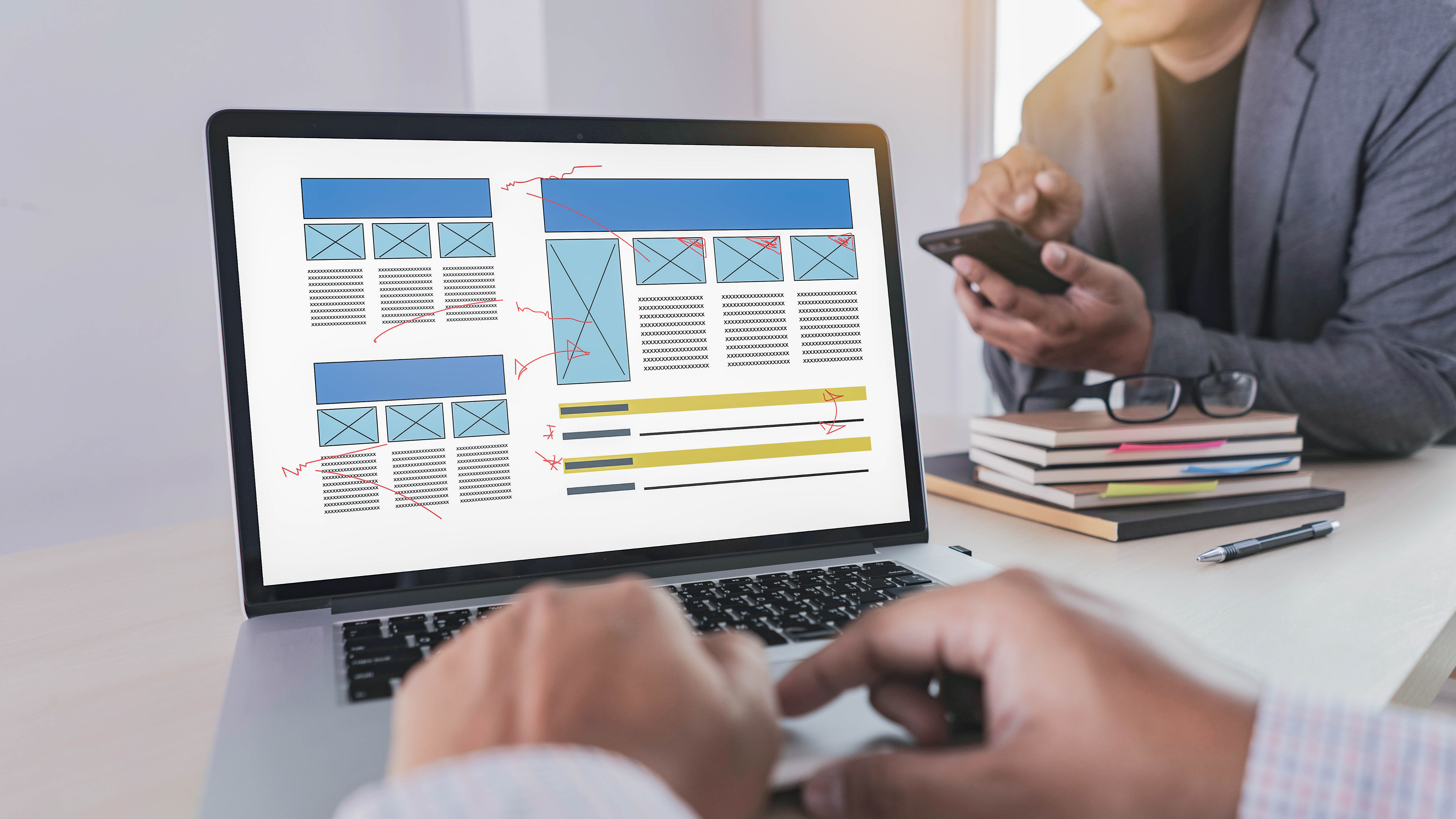 Auf dem Laptop-Bildschirm sind Wireframe-Layouts mit Anmerkungen zu sehen, während eine Person ein Smartphone hält.