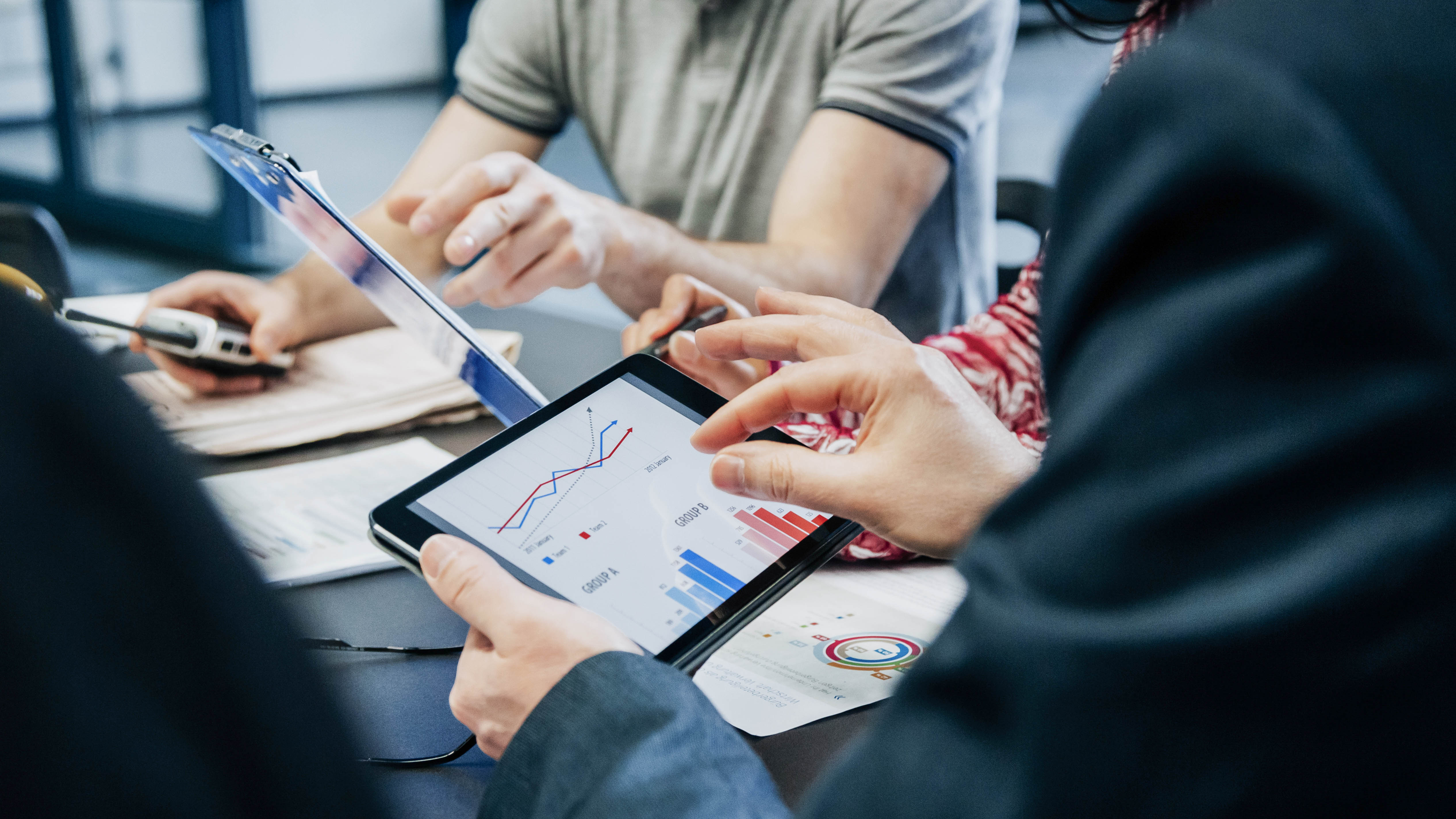 Hände von Personen, die an einem Tisch sitzen und auf ein Tablet mit Diagrammen und Grafiken schauen.