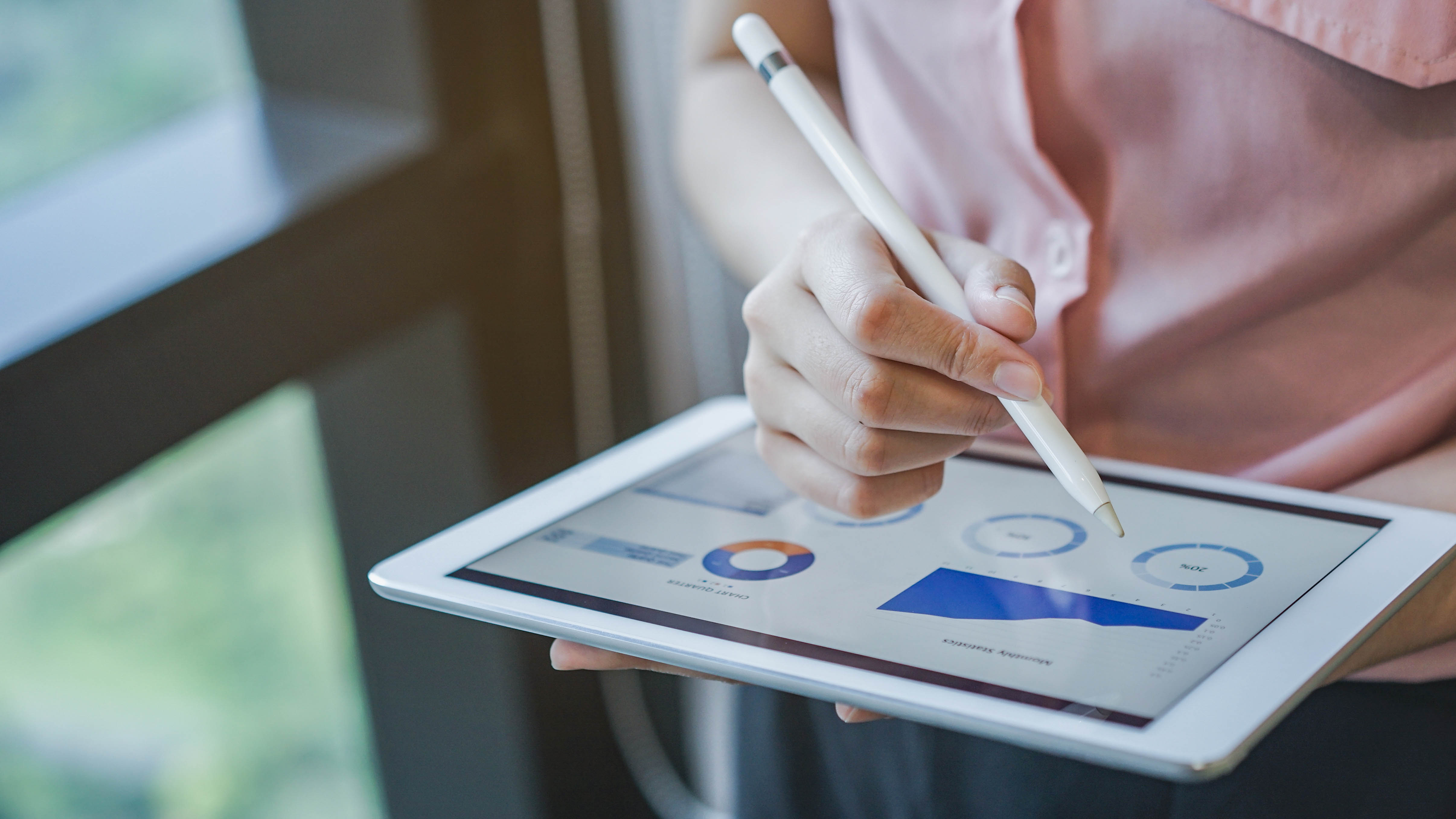 Eine Hand hält ein Tablet, auf dem Diagramme und Grafiken angezeigt werden, während mit einem Stylus darauf geschrieben wird.