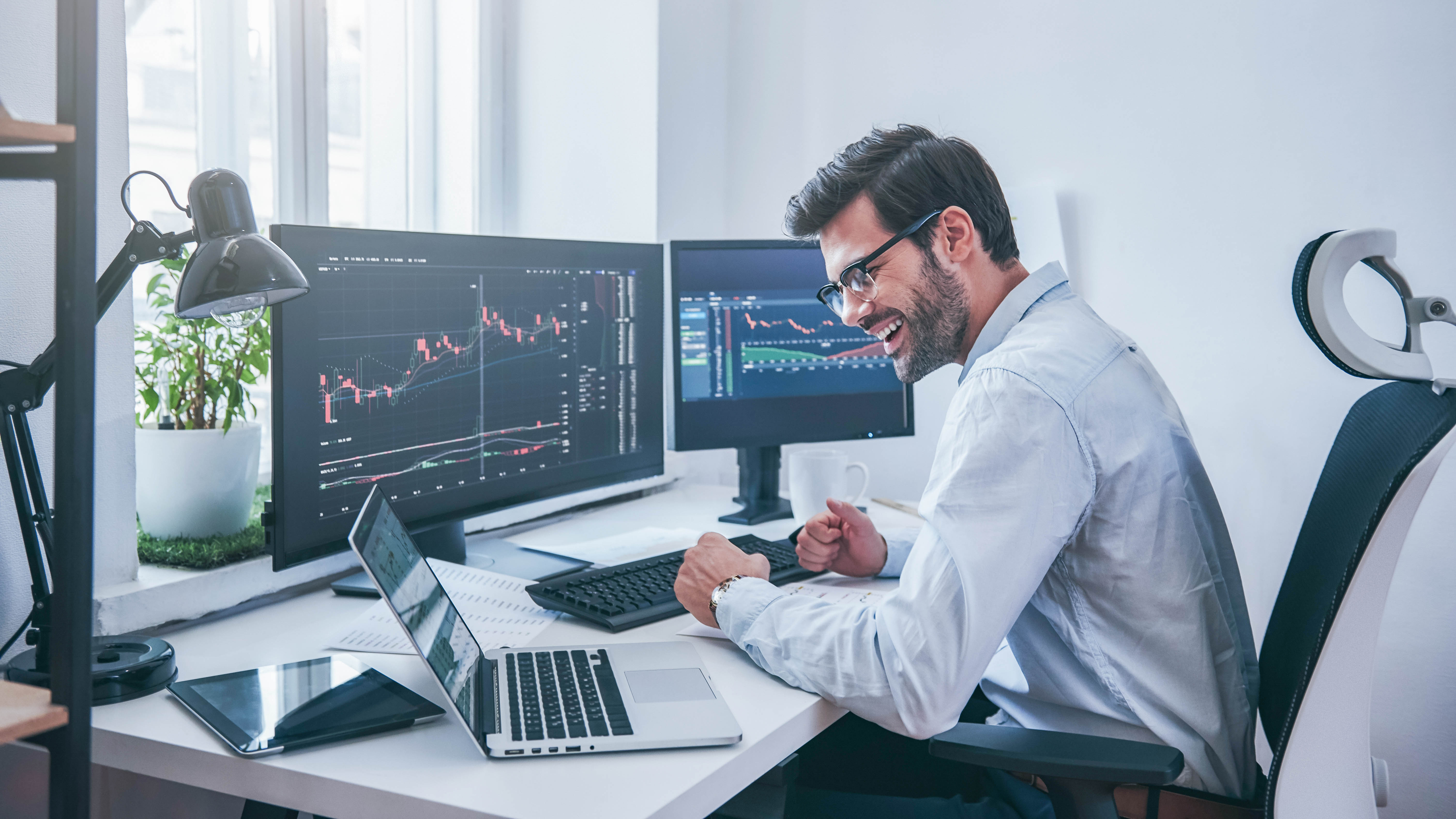 Mann in einem modernen Büro lächelt und feiert vor mehreren Bildschirmen mit Finanzgrafiken.