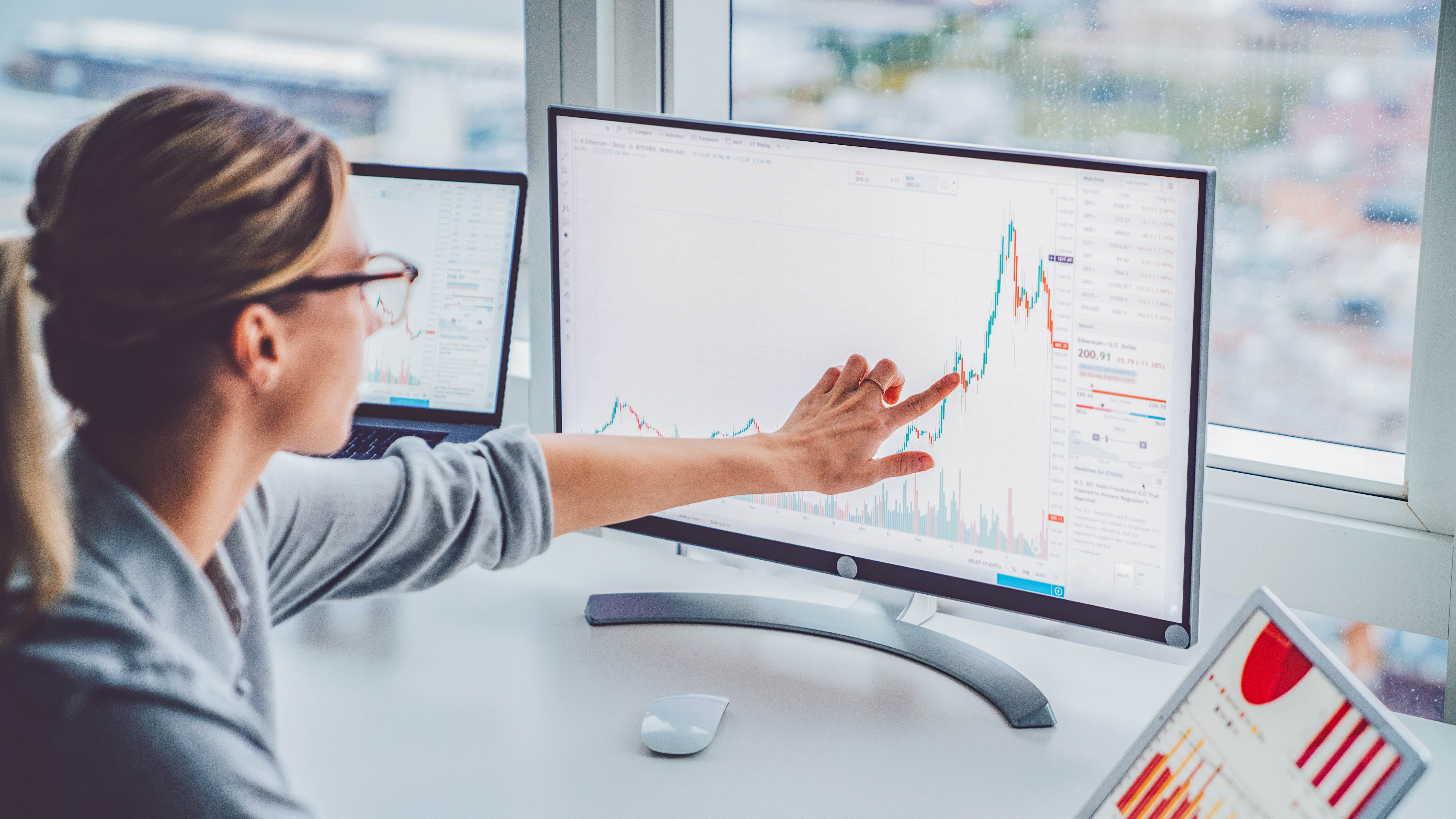 Eine Person zeigt mit dem Finger auf ein Diagramm auf einem Monitor, während ein Laptop und ein weiteres Display im Hintergrund sichtbar sind.
