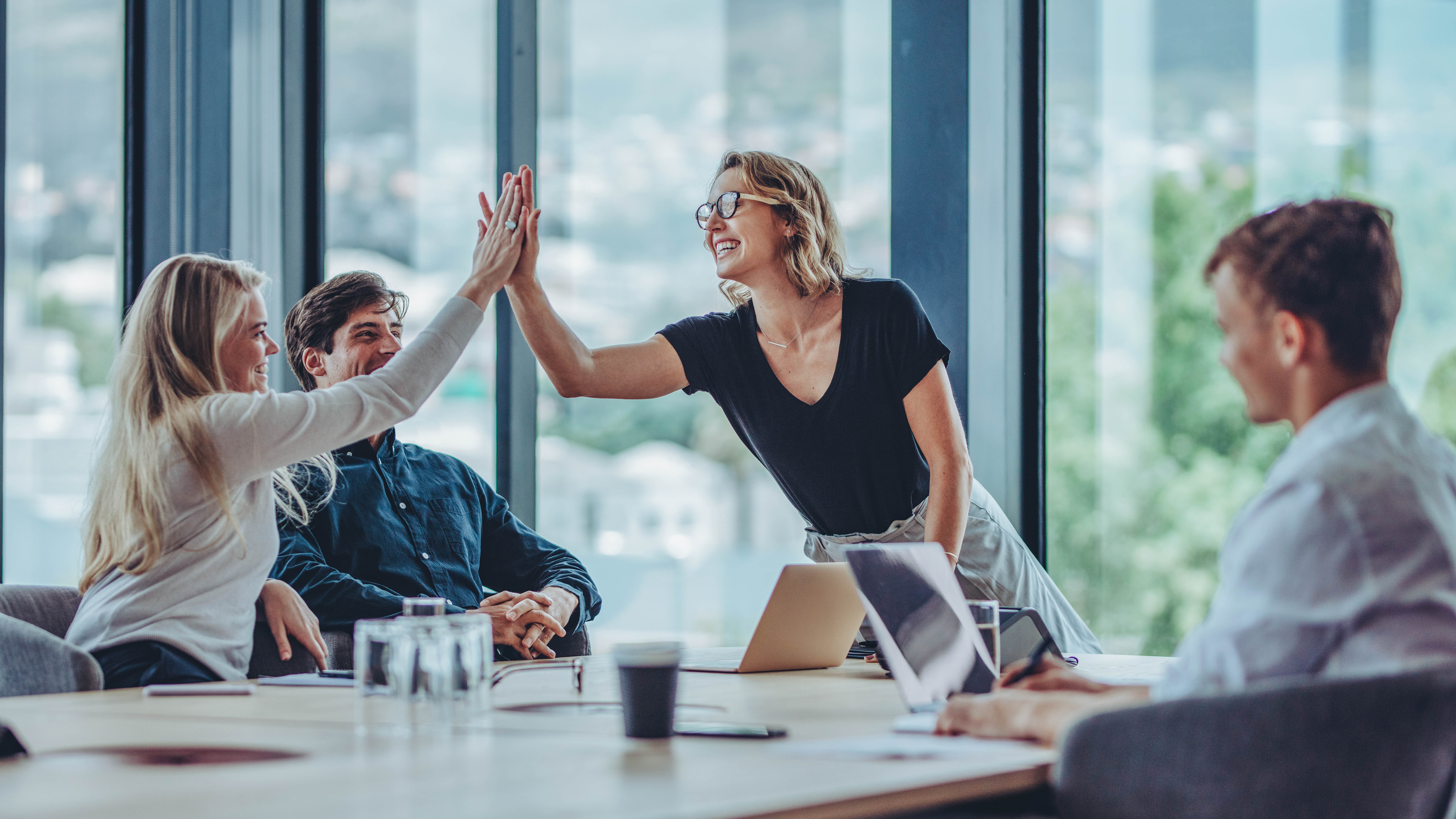 Zwei Personen geben sich in einem modernen Besprechungsraum ein High-Five, während zwei weitere Personen am Tisch sitzen.