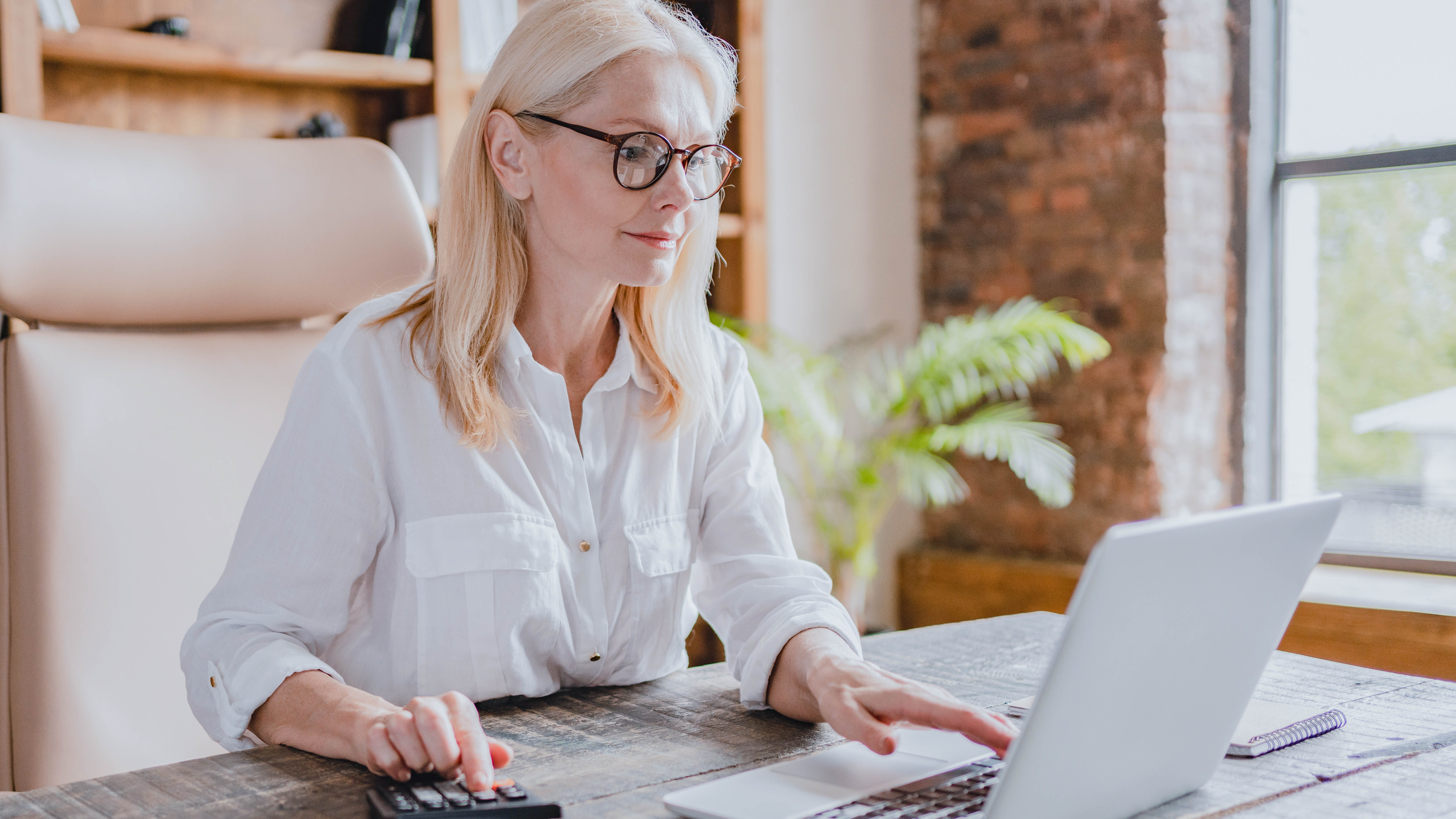 Eine Frau mit blonden Haaren und Brille arbeitet an einem Laptop und benutzt einen Taschenrechner.