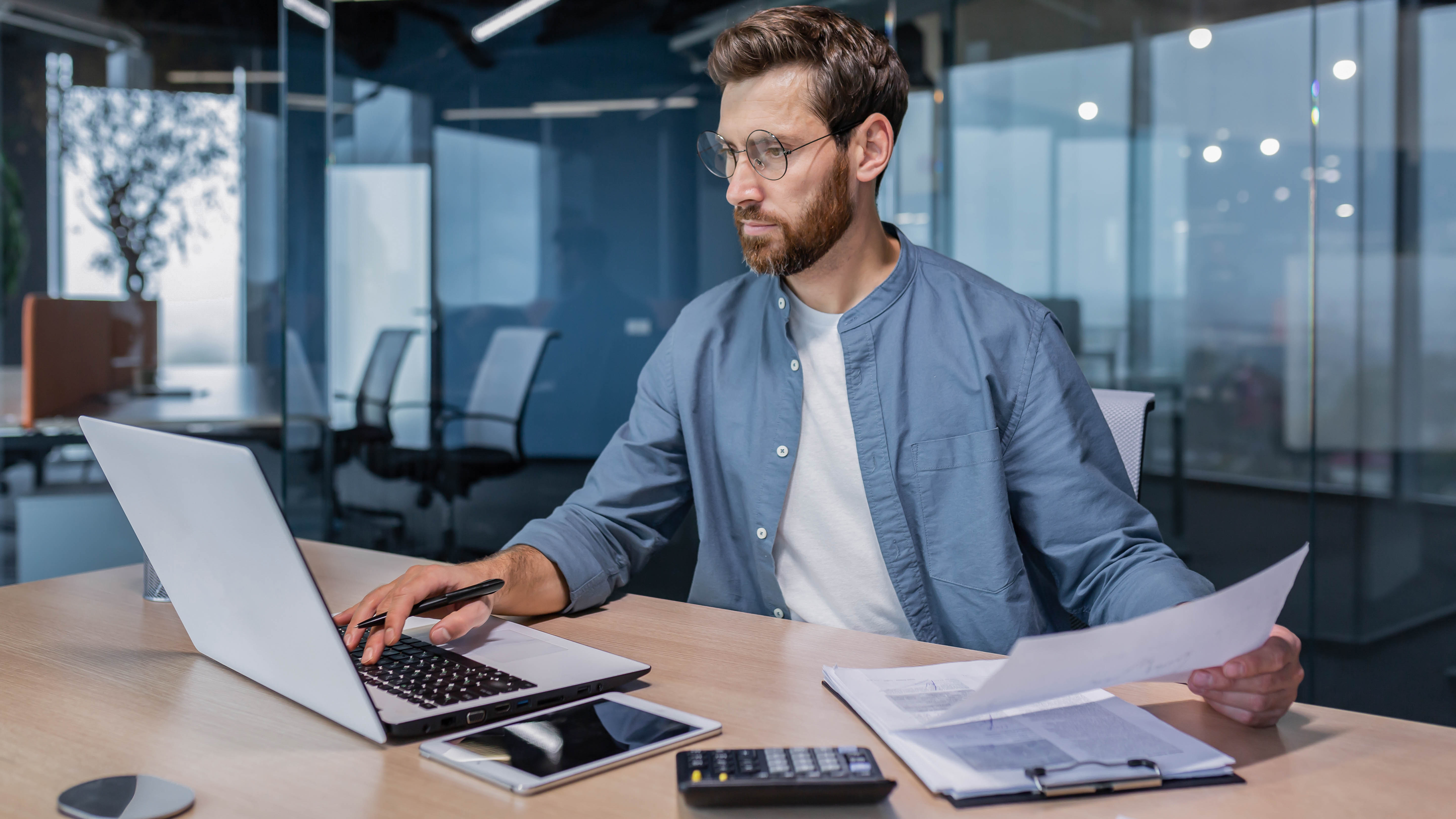 Mann mit Brille sitzt an einem Schreibtisch, arbeitet am Laptop und hält Dokumente in der Hand.