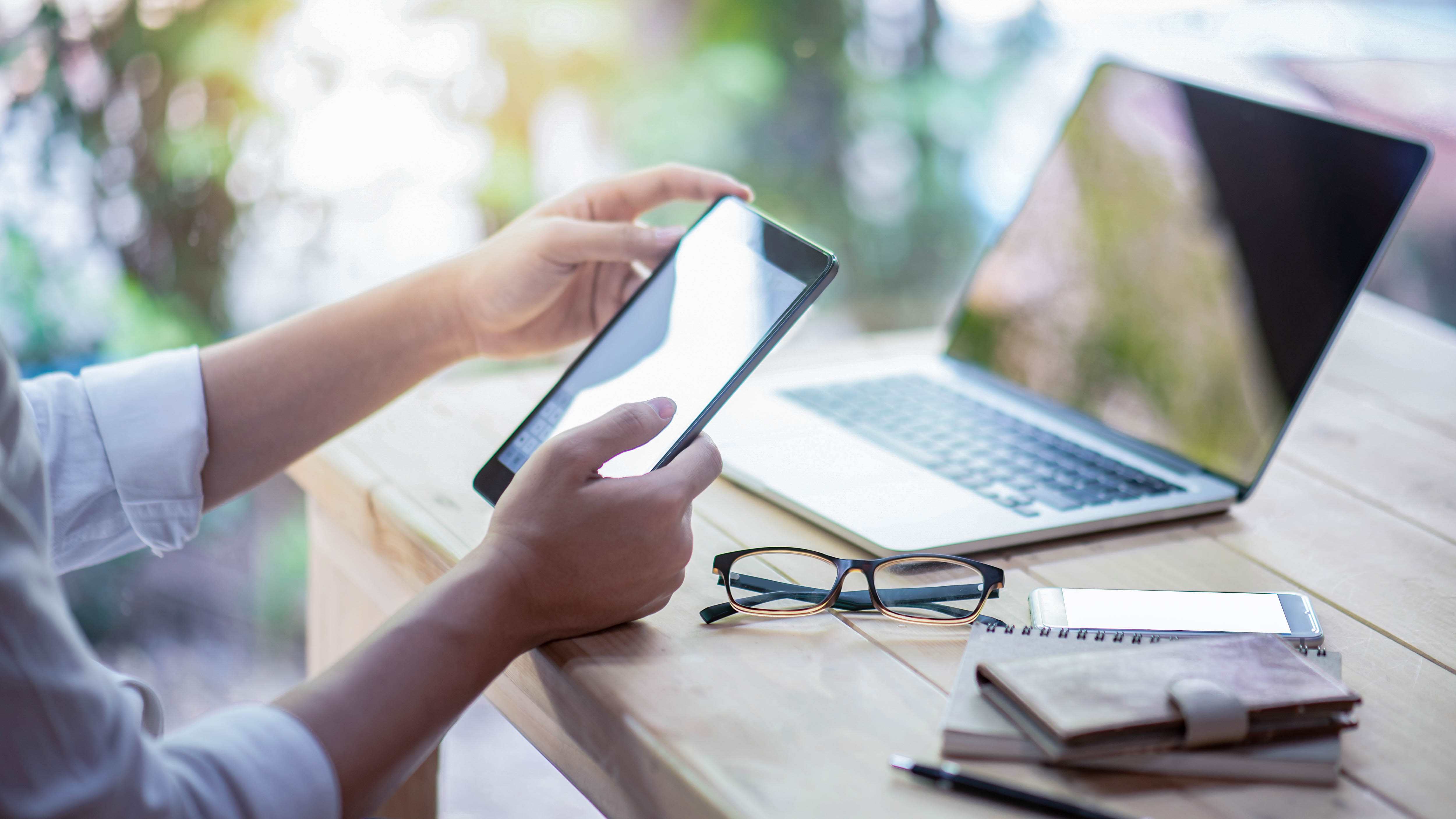 Eine Hand hält ein Tablet, daneben ein Laptop, eine Brille, ein Notizbuch und ein Smartphone auf einem Tisch.