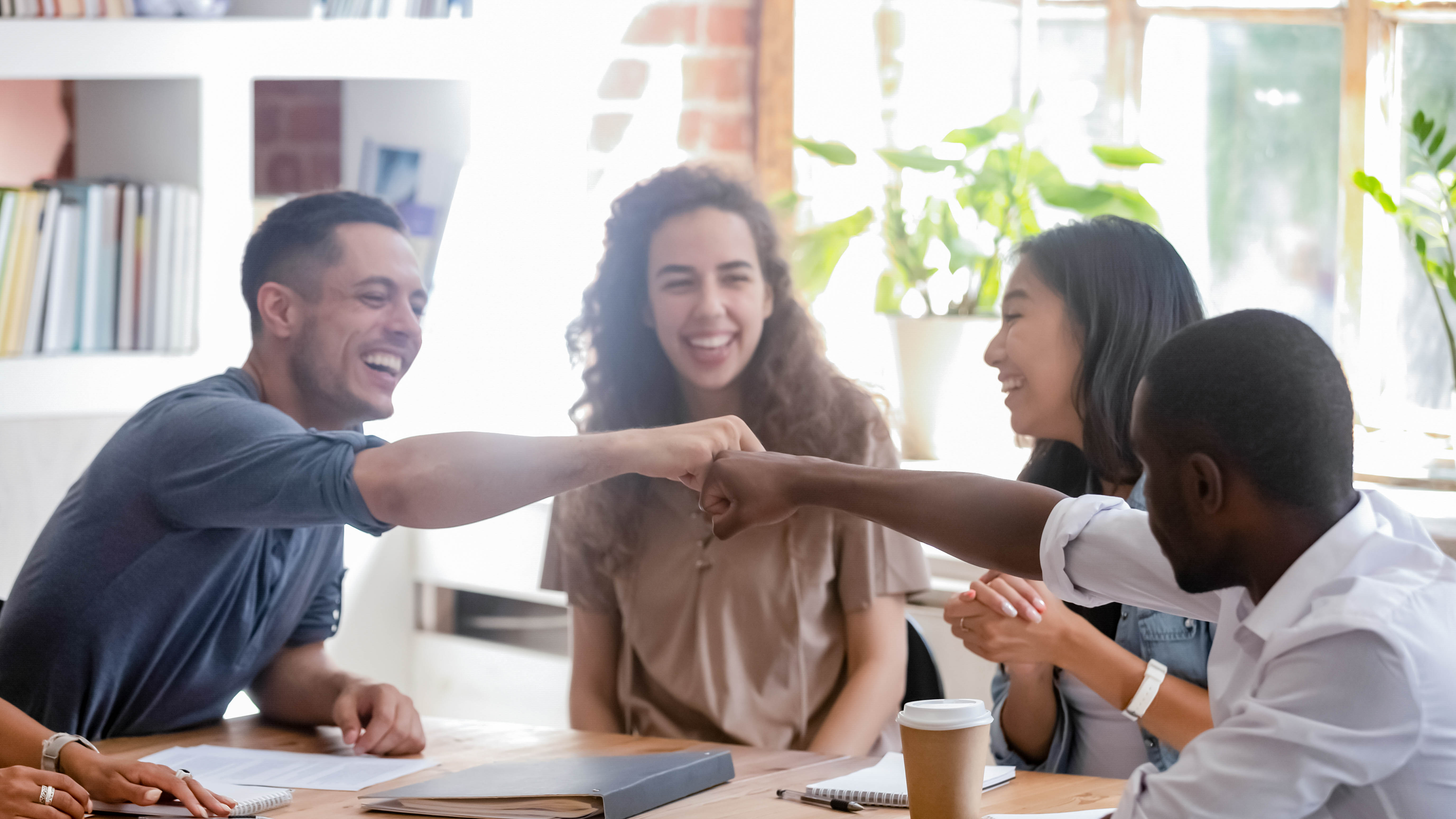 Vier Personen sitzen an einem Tisch und lachen, während sie sich mit einem Fistbump begrüßen.