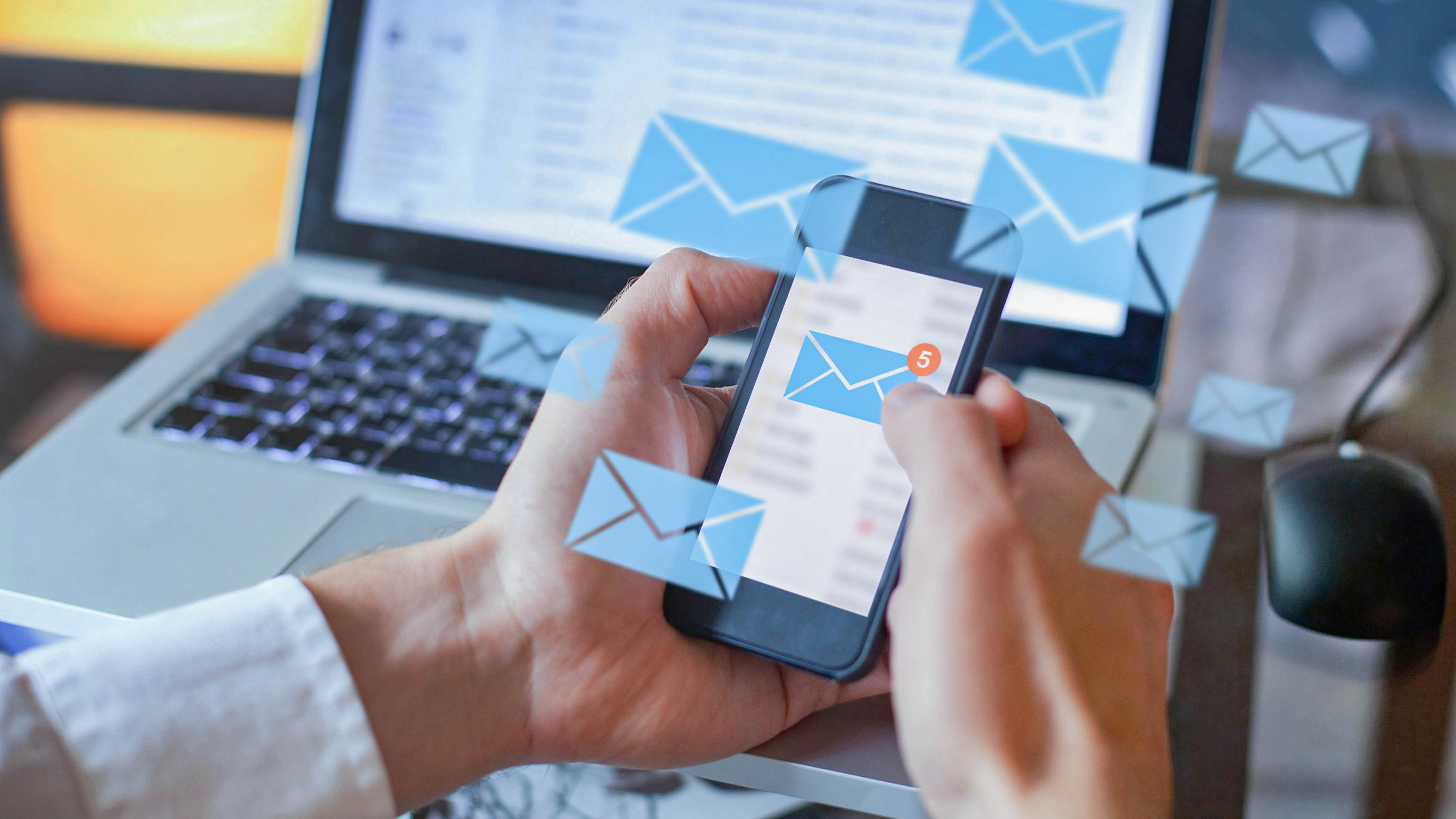 Eine Person hält ein Smartphone in der Hand, auf dem Benachrichtigungen für neue E-Mails angezeigt werden, während ein Laptop im Hintergrund sichtbar ist.