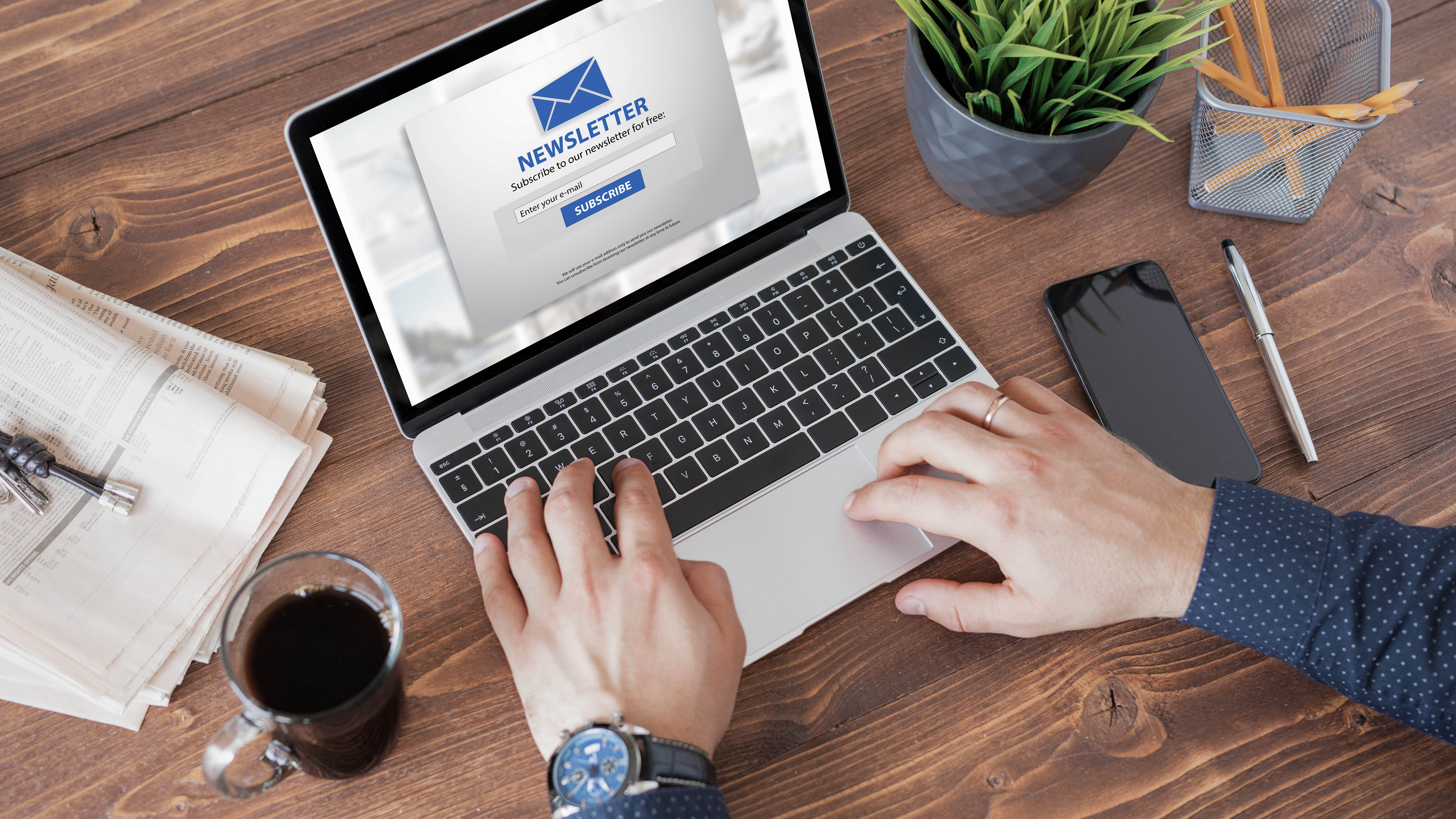 Hände tippen auf einem Laptop, der eine Newsletter-Anmeldeseite zeigt, umgeben von einer Tasse Kaffee und Büromaterial.