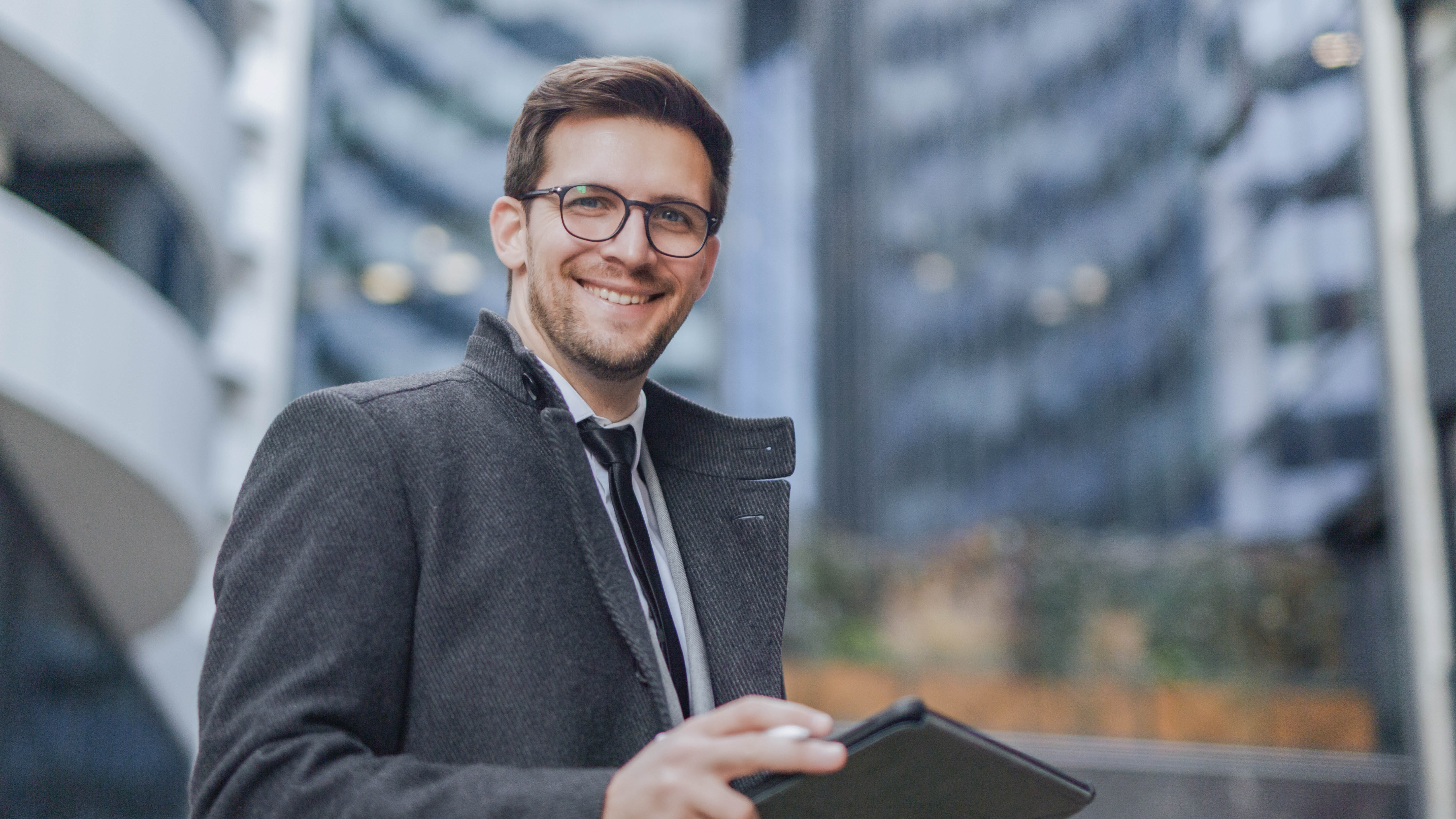 Lächelnder Mann in einem grauen Mantel hält ein Tablet in der Hand, vor modernen Gebäuden im Hintergrund.