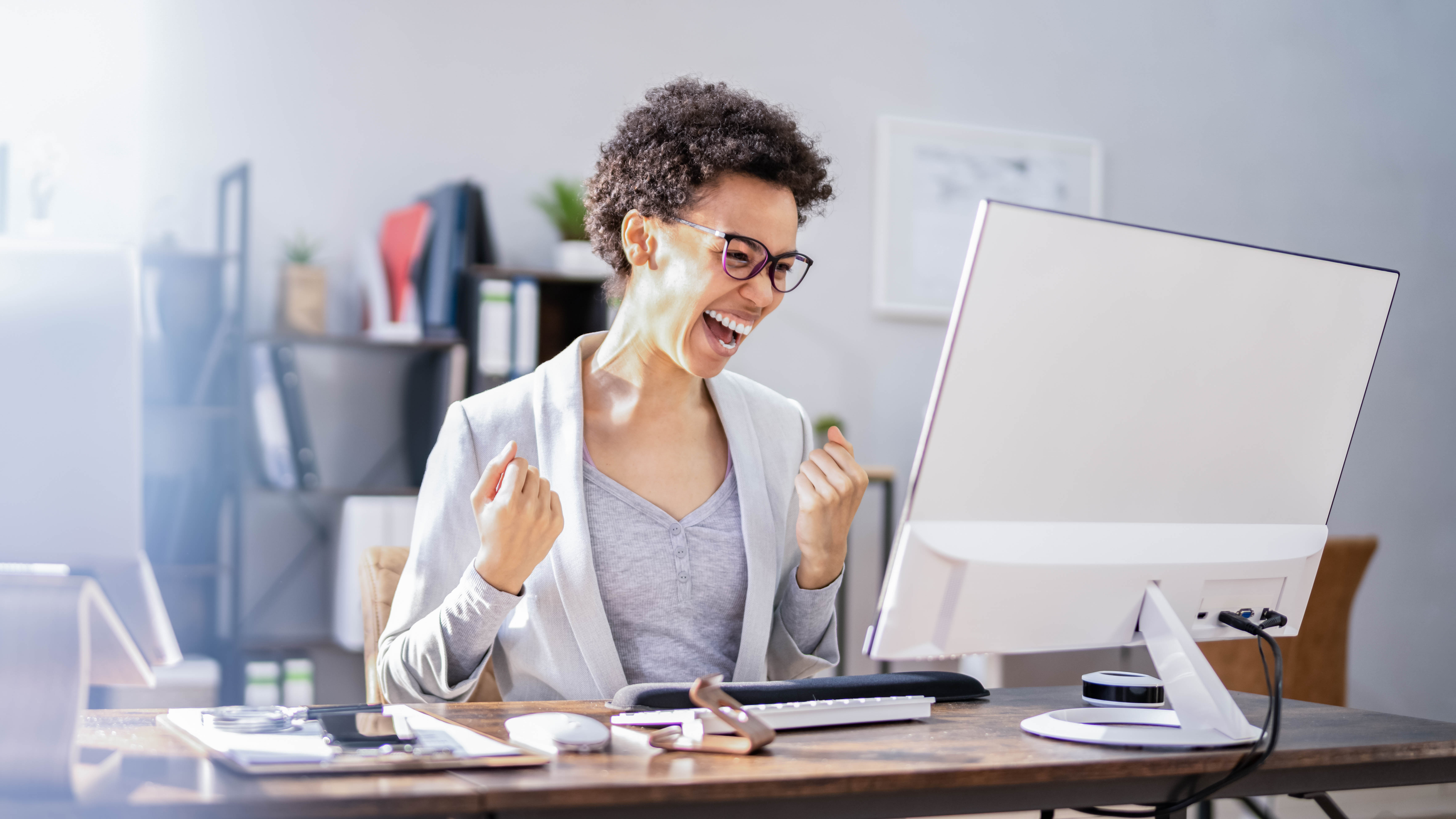 Frau mit lockigem Haar und Brille sitzt am Schreibtisch, lächelt und jubelt vor einem Computerbildschirm.