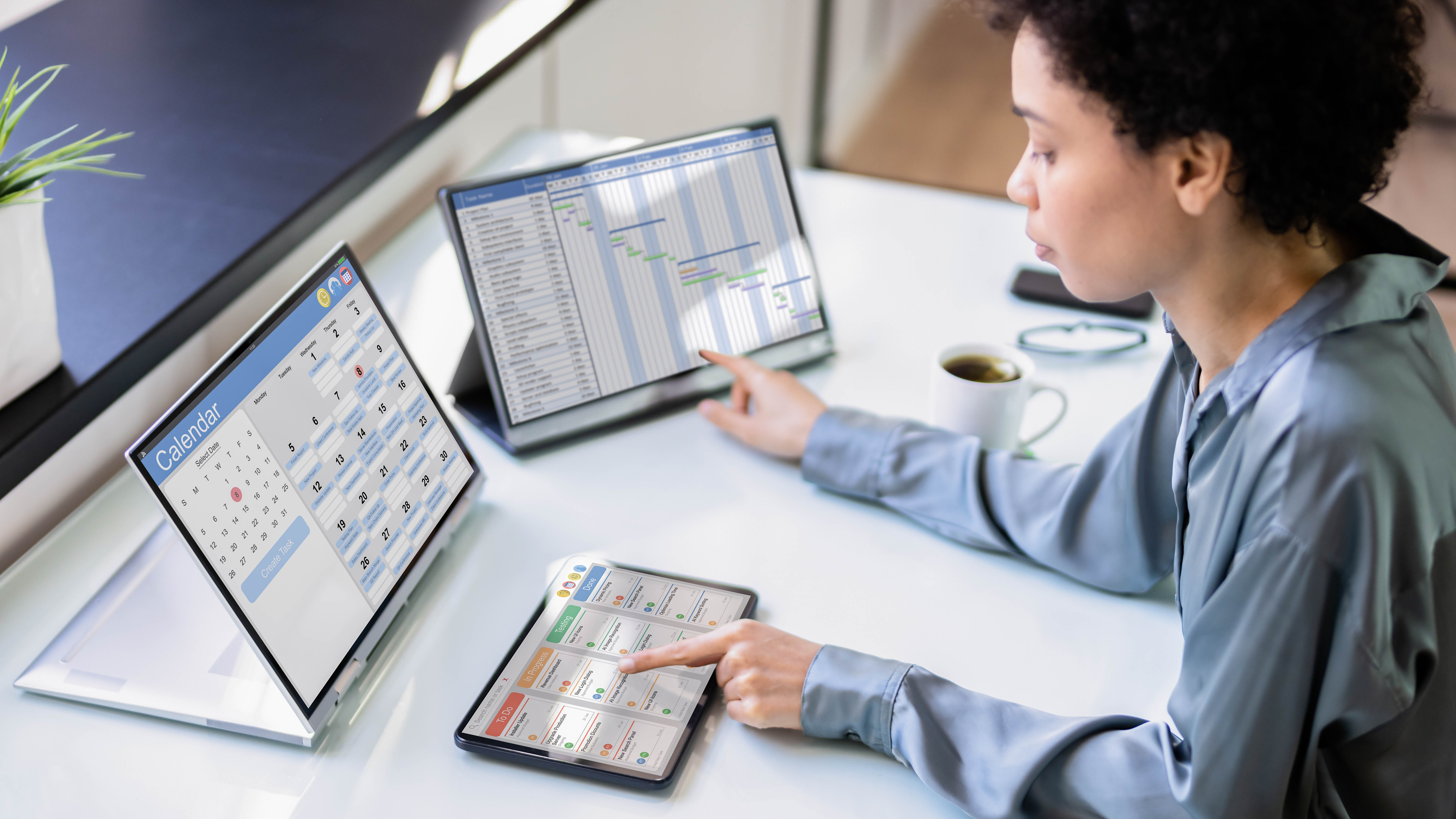 Eine Person mit lockigem Haar sitzt an einem Tisch und nutzt ein Tablet sowie zwei Laptops mit Kalender- und Projektmanagement-Software.