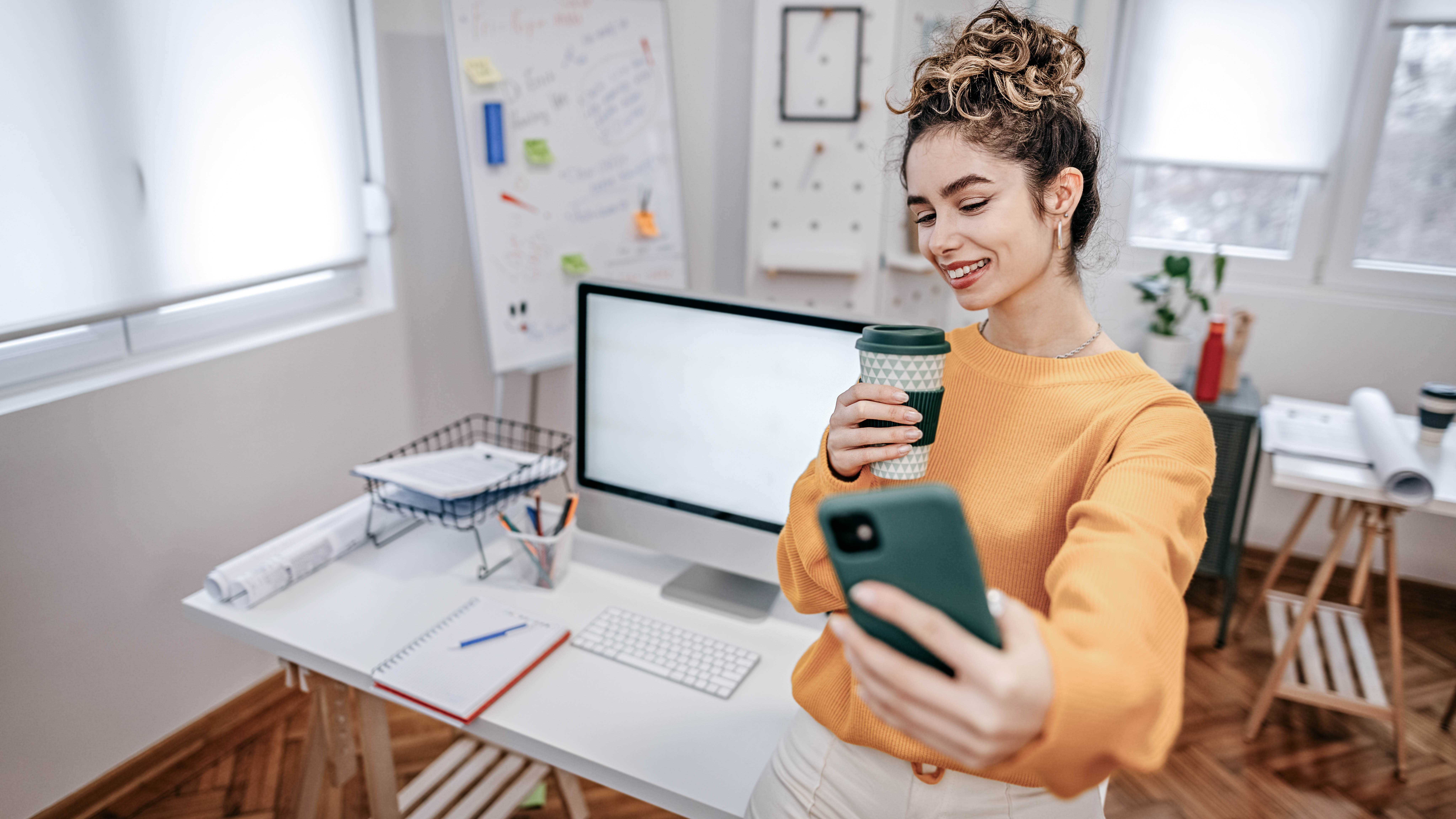 Eine lächelnde Frau in einem orangefarbenen Pullover macht ein Selfie, während sie einen Kaffeebecher hält.