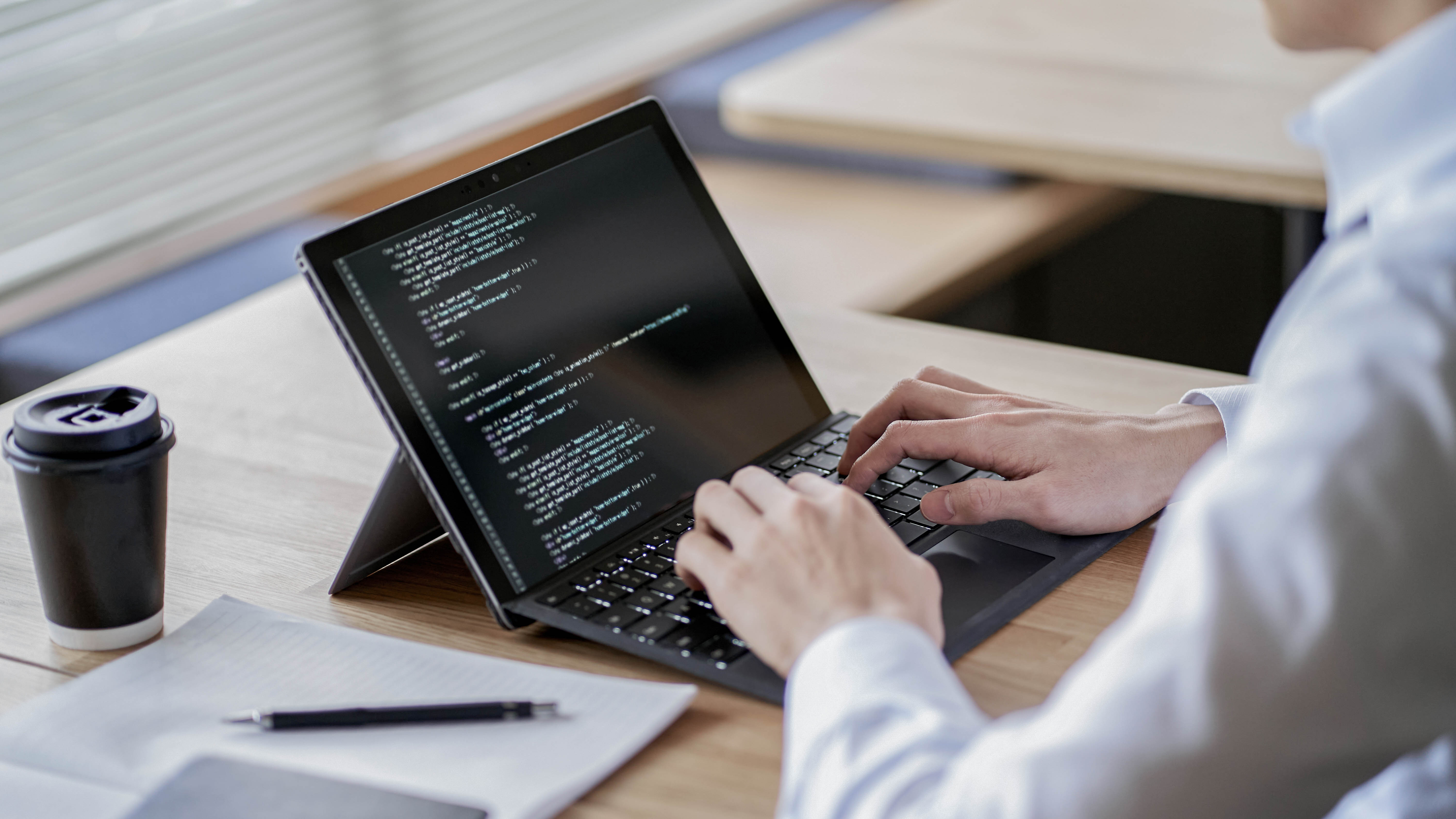Eine Person tippt auf einem Laptop mit Code auf dem Bildschirm, daneben ein Kaffeebecher und ein Notizblock.