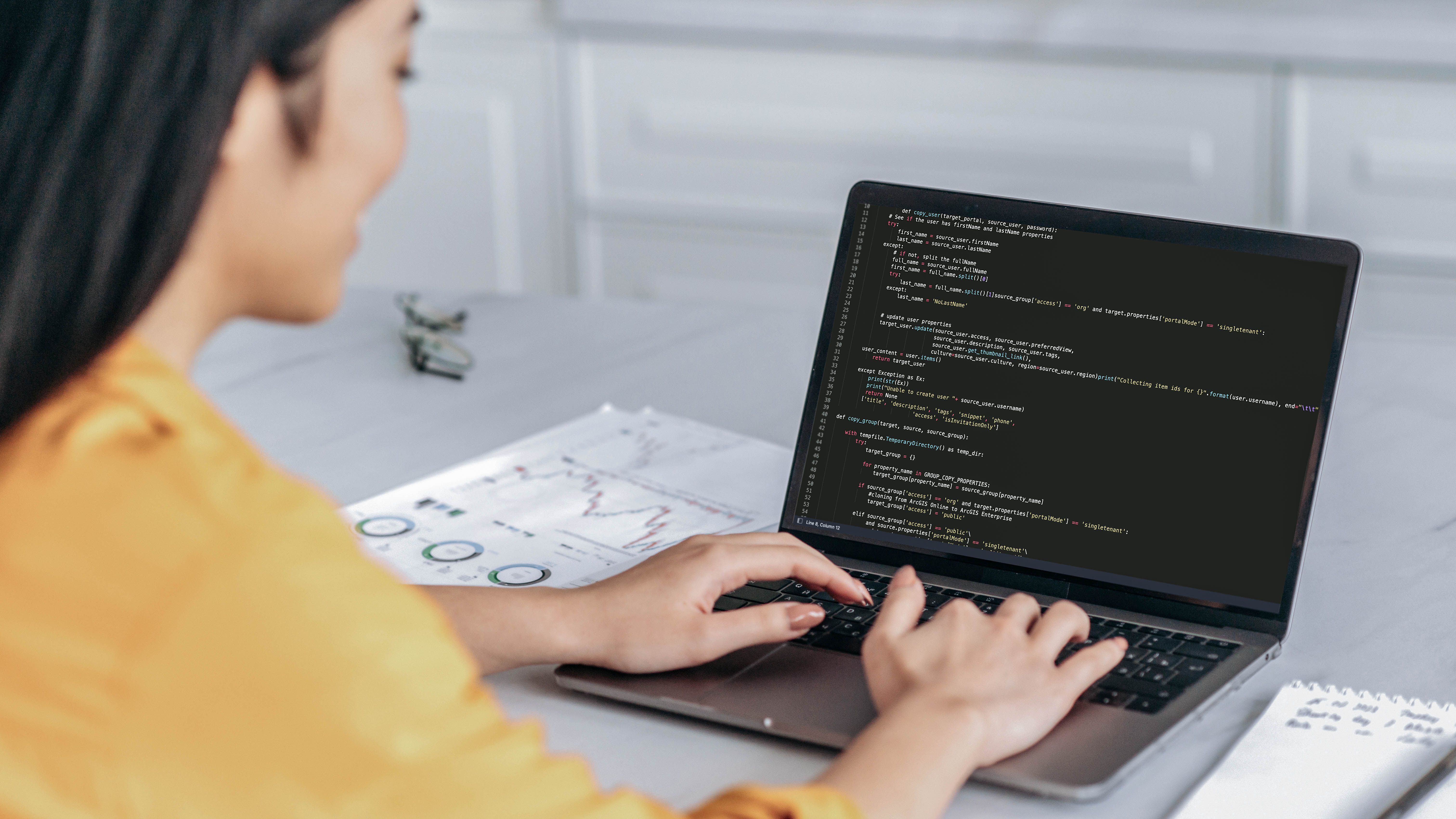 Eine Person mit langen Haaren tippt auf einem Laptop, auf dem Programmiercode sichtbar ist. Im Hintergrund liegen Diagramme und Notizen.