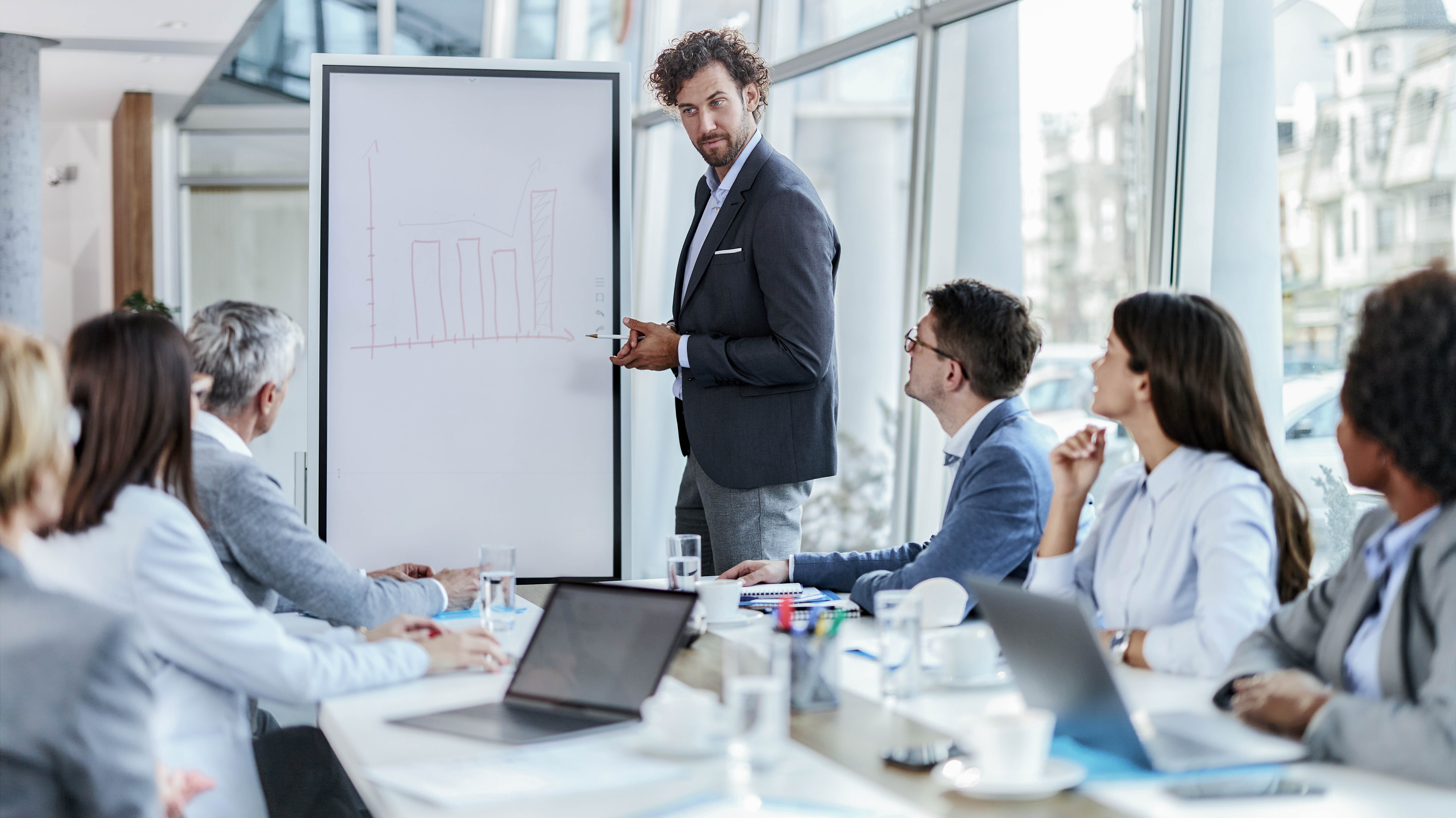 Ein Mann präsentiert an einem Flipchart mit Diagramm vor einer Gruppe von Personen in einem Konferenzraum.