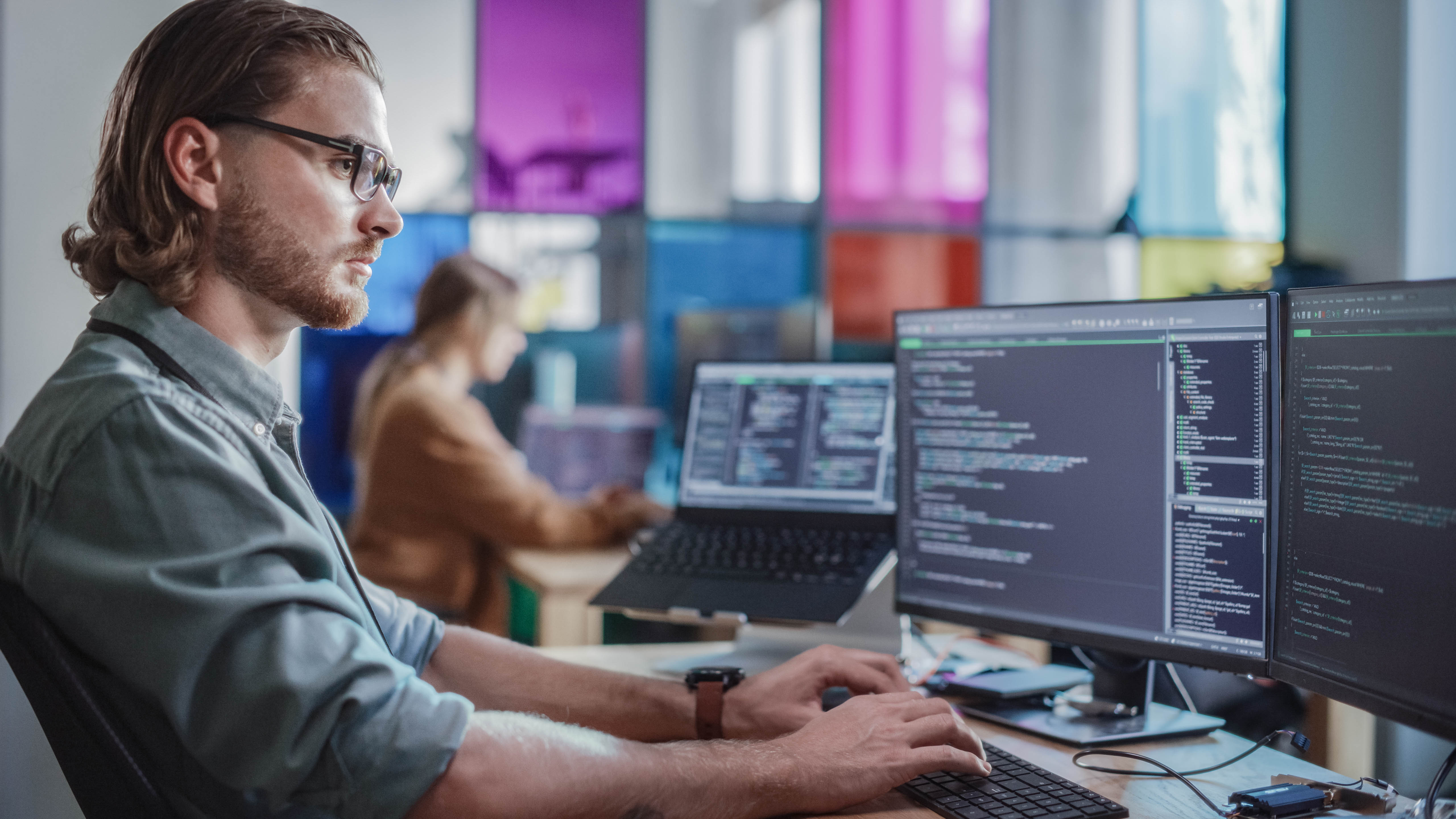 Mann mit Brille arbeitet an zwei Monitoren, die Programmiercode anzeigen, im modernen Büro mit bunten Fenstern.